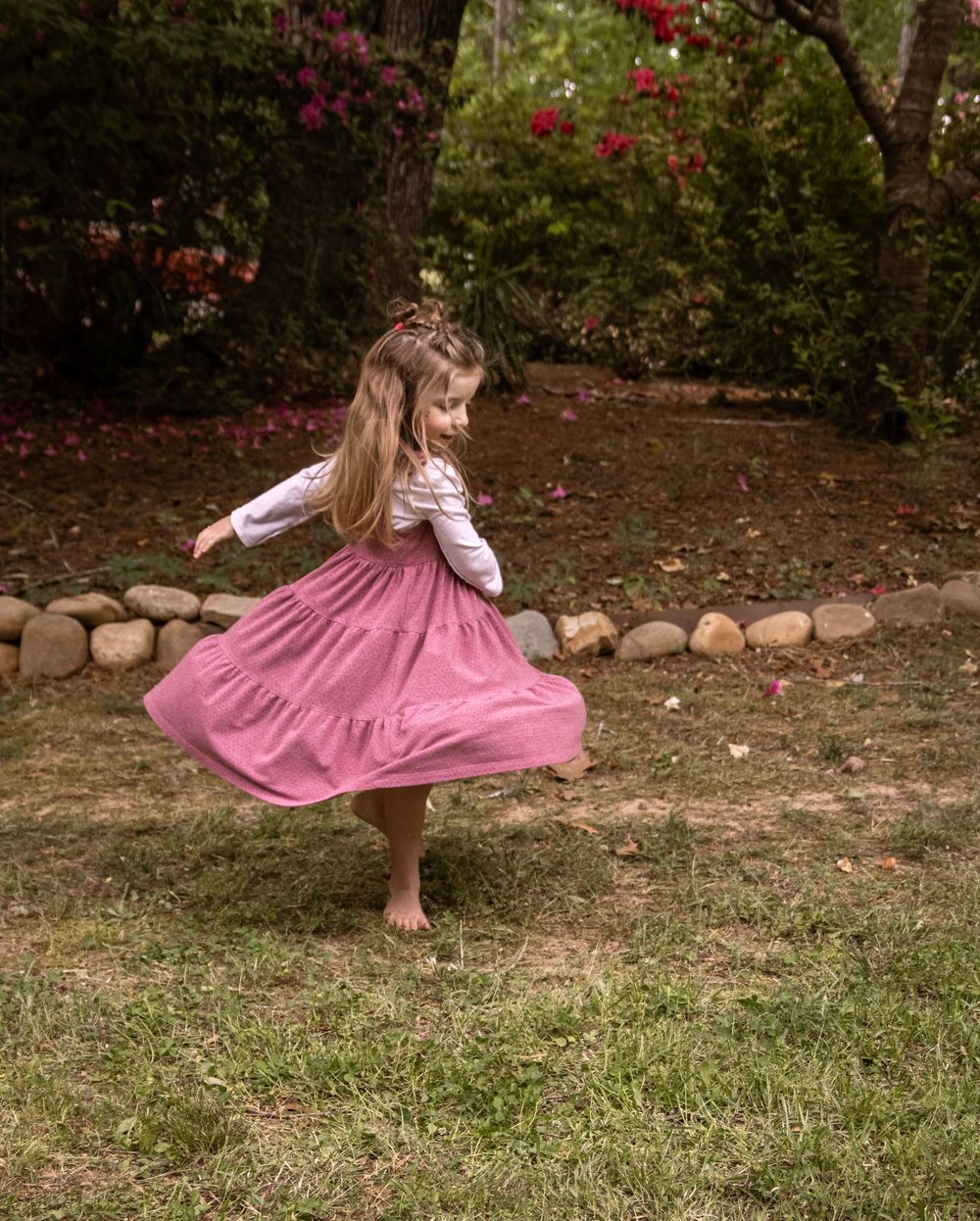 Young girl in pink tiered dress twirling barefoot in a garden with colorful flowers and stones.