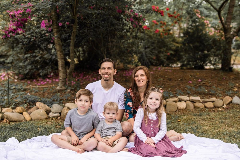 Happy family of five sitting on white blanket in garden with blooming pink flowers and stone wall.