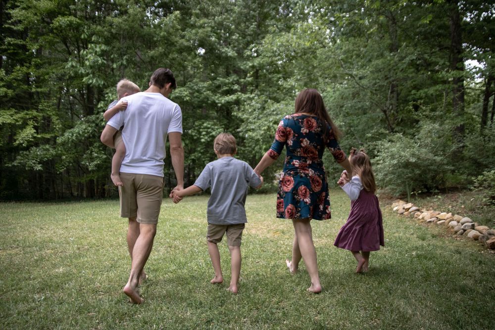 Family of five walking hand-in-hand across green lawn toward lush forest, viewed from behind.