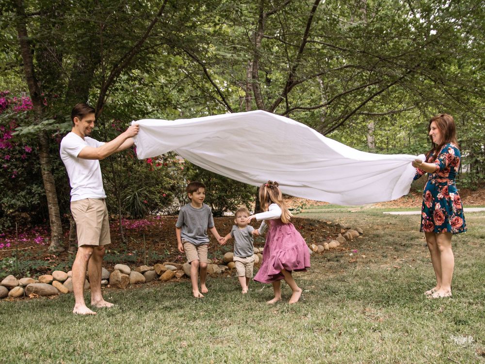 Family playing with a white sheet outdoors in a lush green backyard with children running underneath.