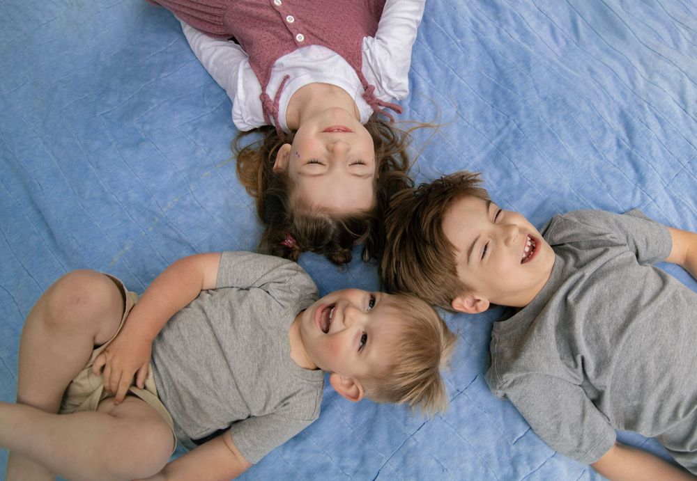 Three happy children lying on a blue blanket together, smiling and laughing from above.