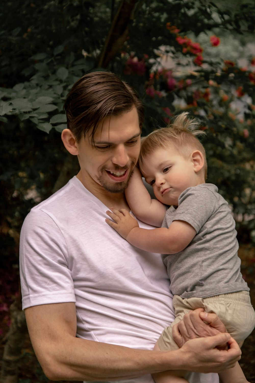 A young father smiling while holding his toddler son outdoors, with lush green and red flowering bushes in the background.