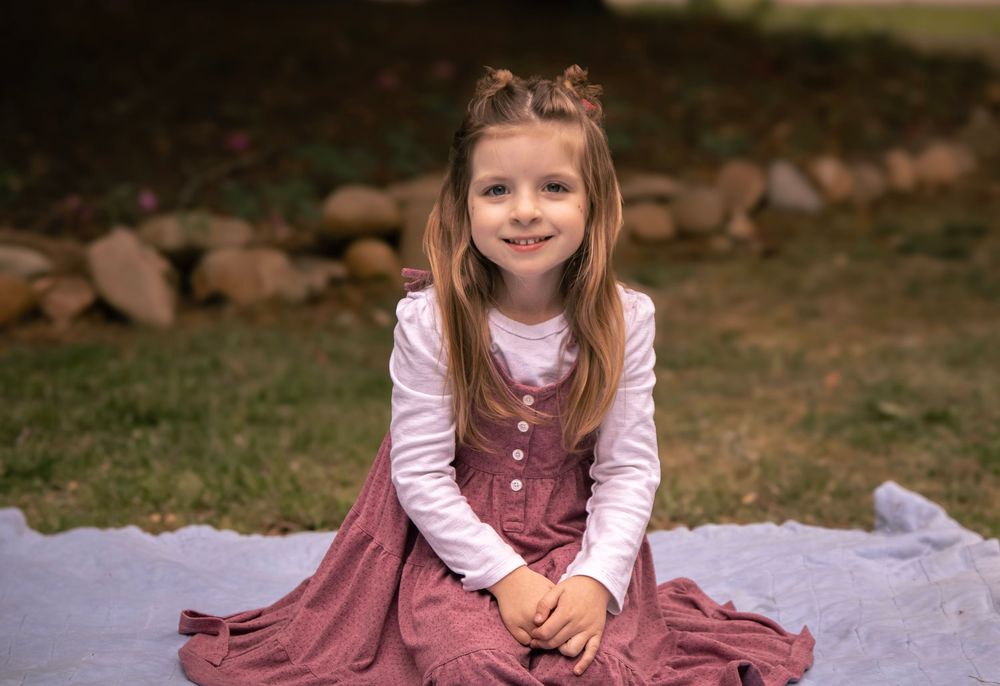 A smiling little girl in a pink dress sits on a blanket outdoors.