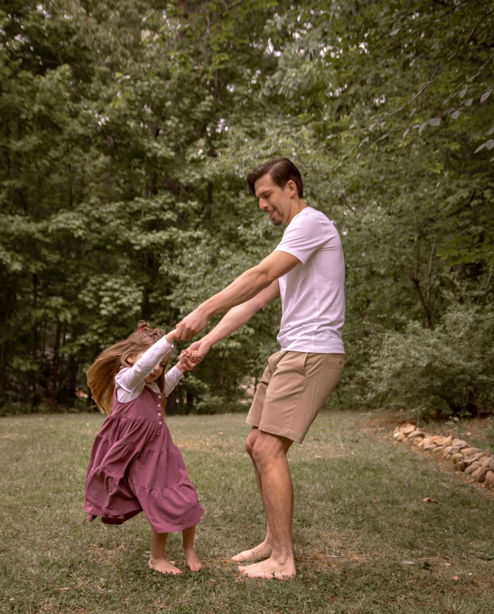 Father spinning young daughter in purple dress outdoors on green lawn surrounded by trees.