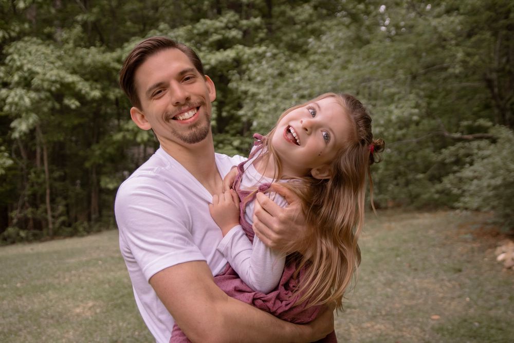 Smiling man in white t-shirt holding a laughing little girl in pink outdoors in a green park.