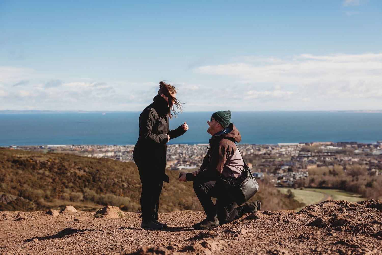Couple sharing a romantic marriage proposal on a hilltop with a scenic coastal city view under a clear blue sky.