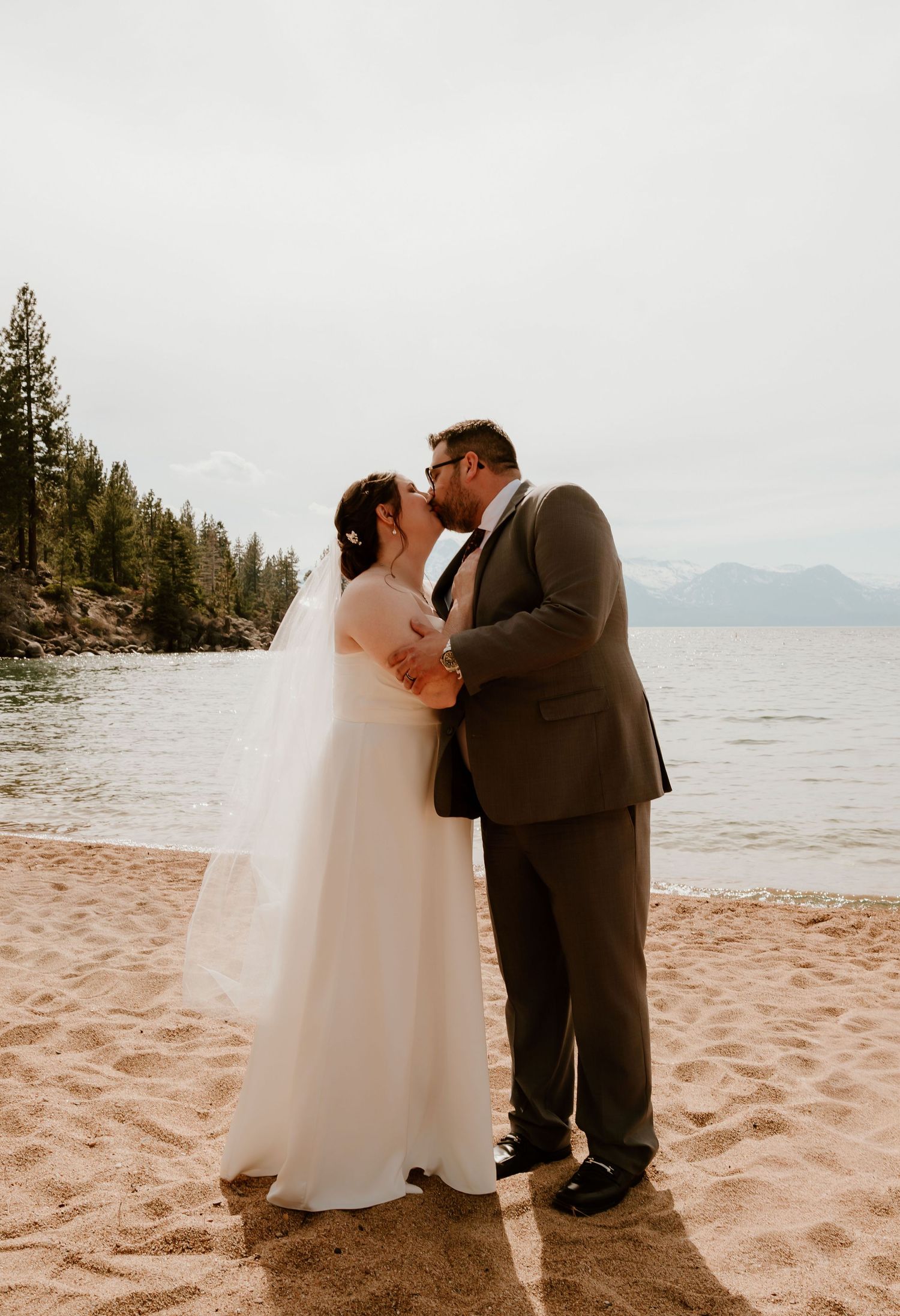 A couple shares a romantic moment on a sandy beach with mountains and evergreen trees in the background at sunset.