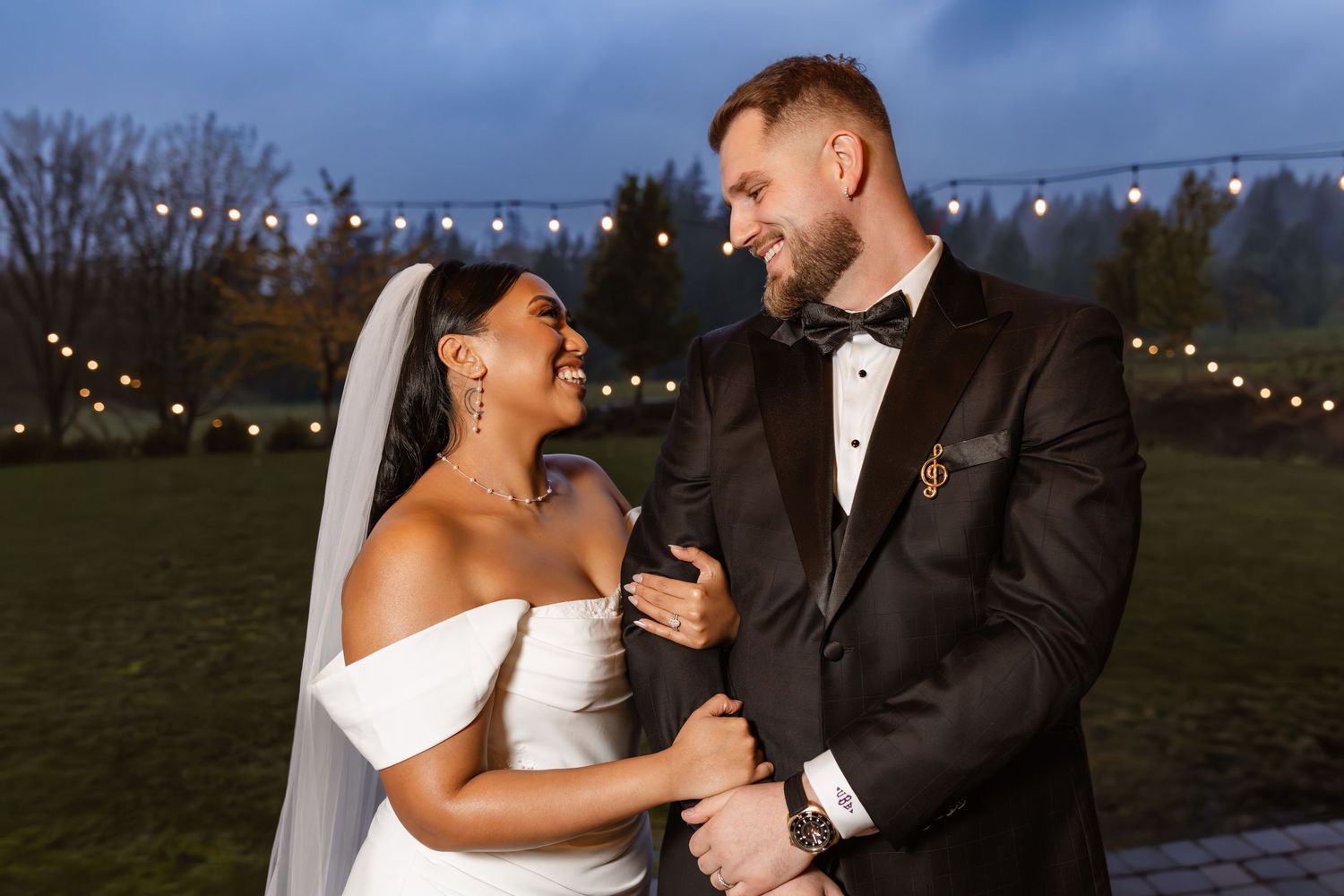 A happy couple shares a joyful moment together outdoors at their evening wedding with string lights in the background.