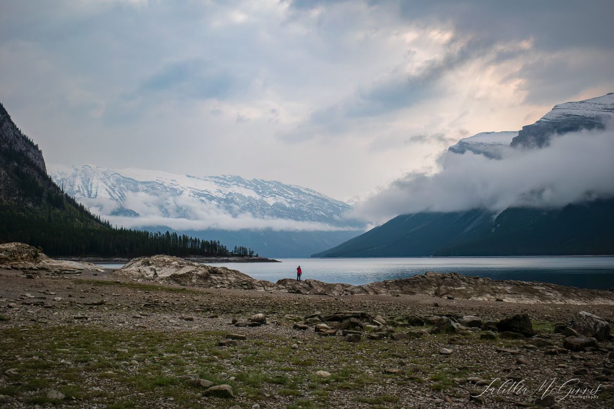 A lone figure stands on the shore of a misty mountain lake surrounded by snow-capped peaks and dramatic clouds.