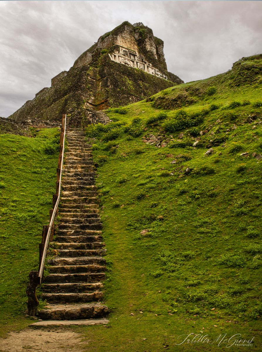 Stone steps ascending a steep green hillside lead to Xunantunich temple on a cloudy day.