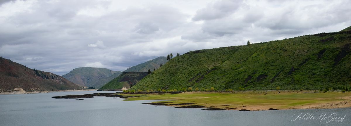 Panoramic view of a mountain reservoir with green hills and cloudy skies reflecting on calm waters.