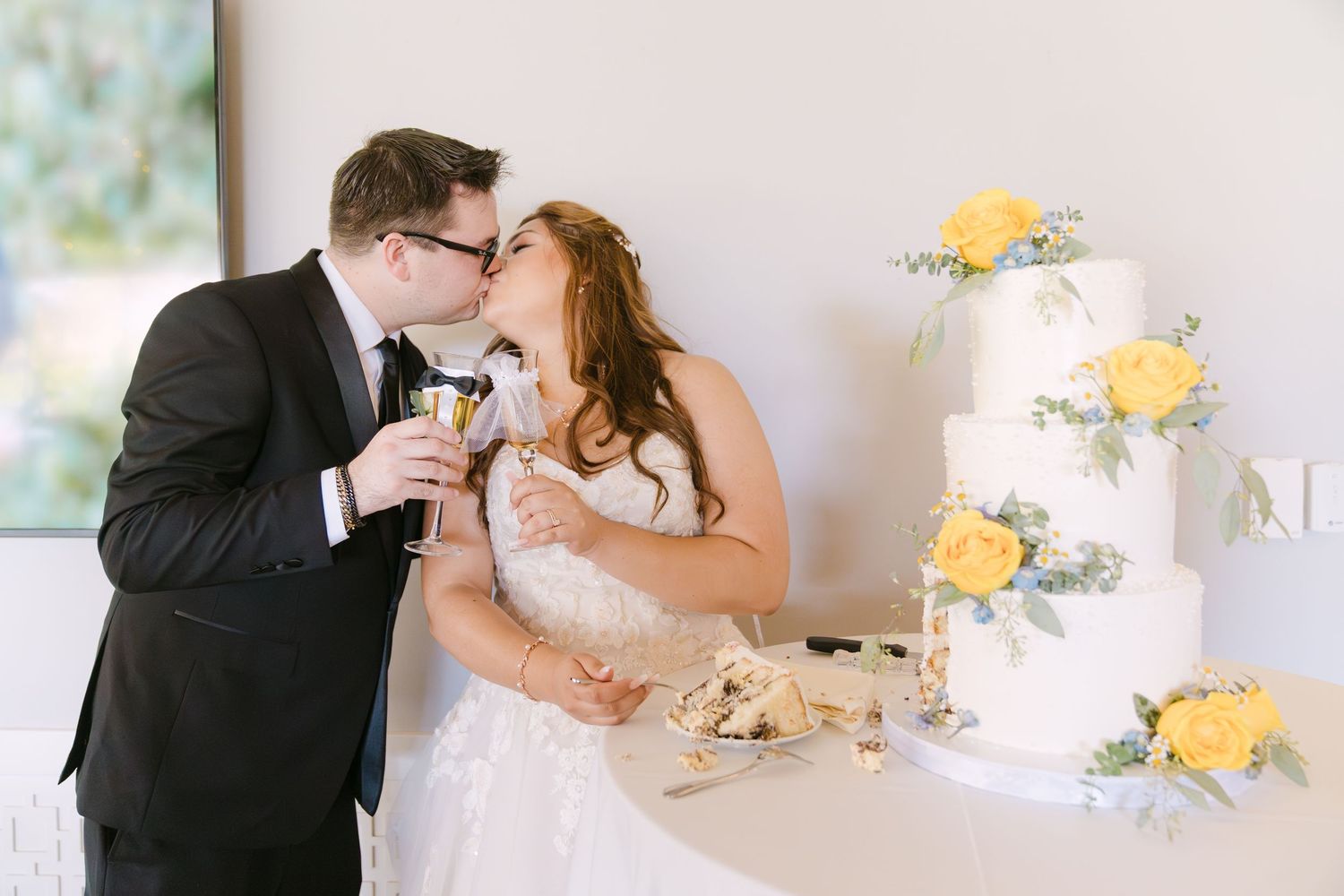 A couple shares a kiss while toasting champagne next to their elegant white wedding cake decorated with yellow flowers.