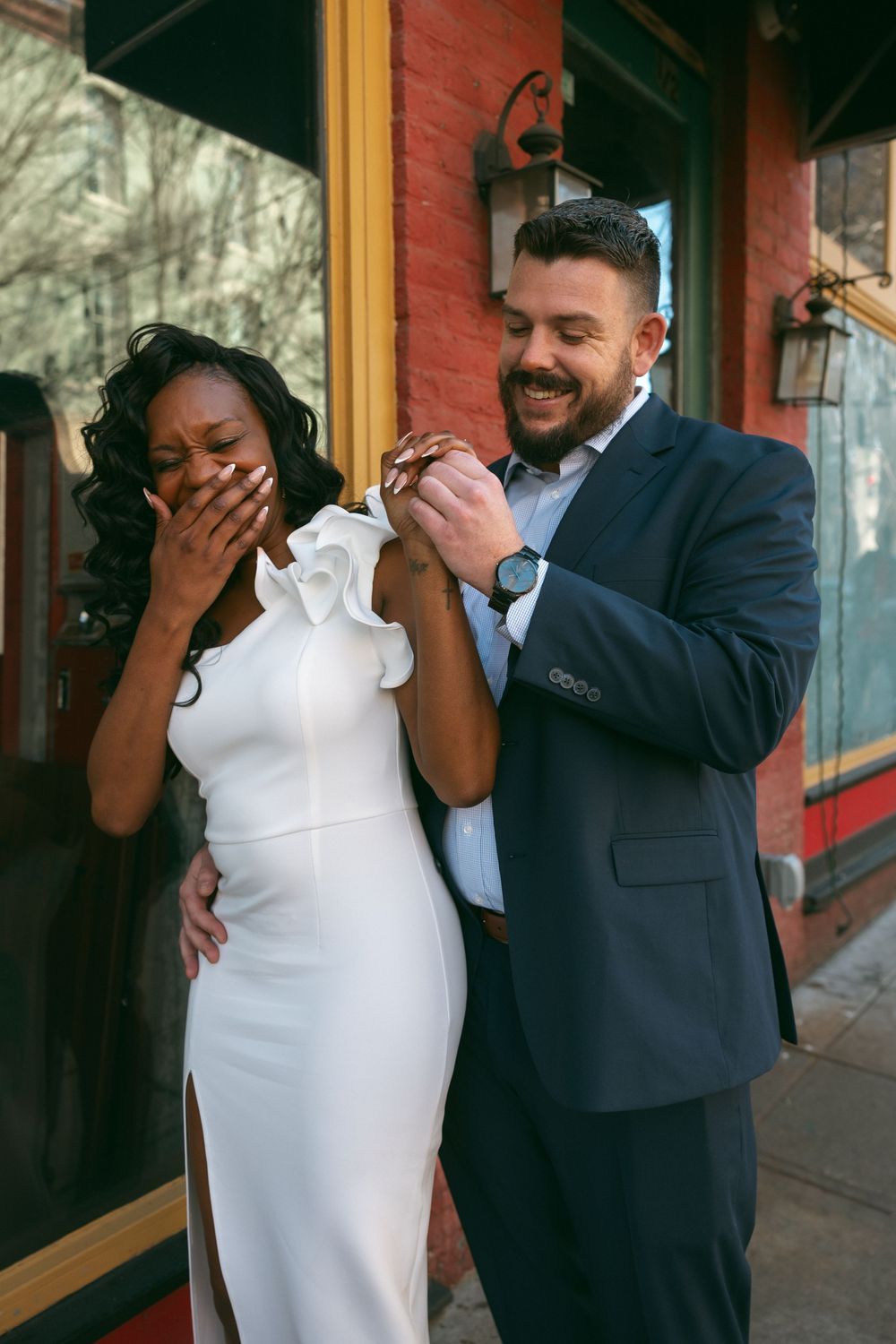 A joyful couple in formal attire share an emotional moment outside a building with a red brick wall.