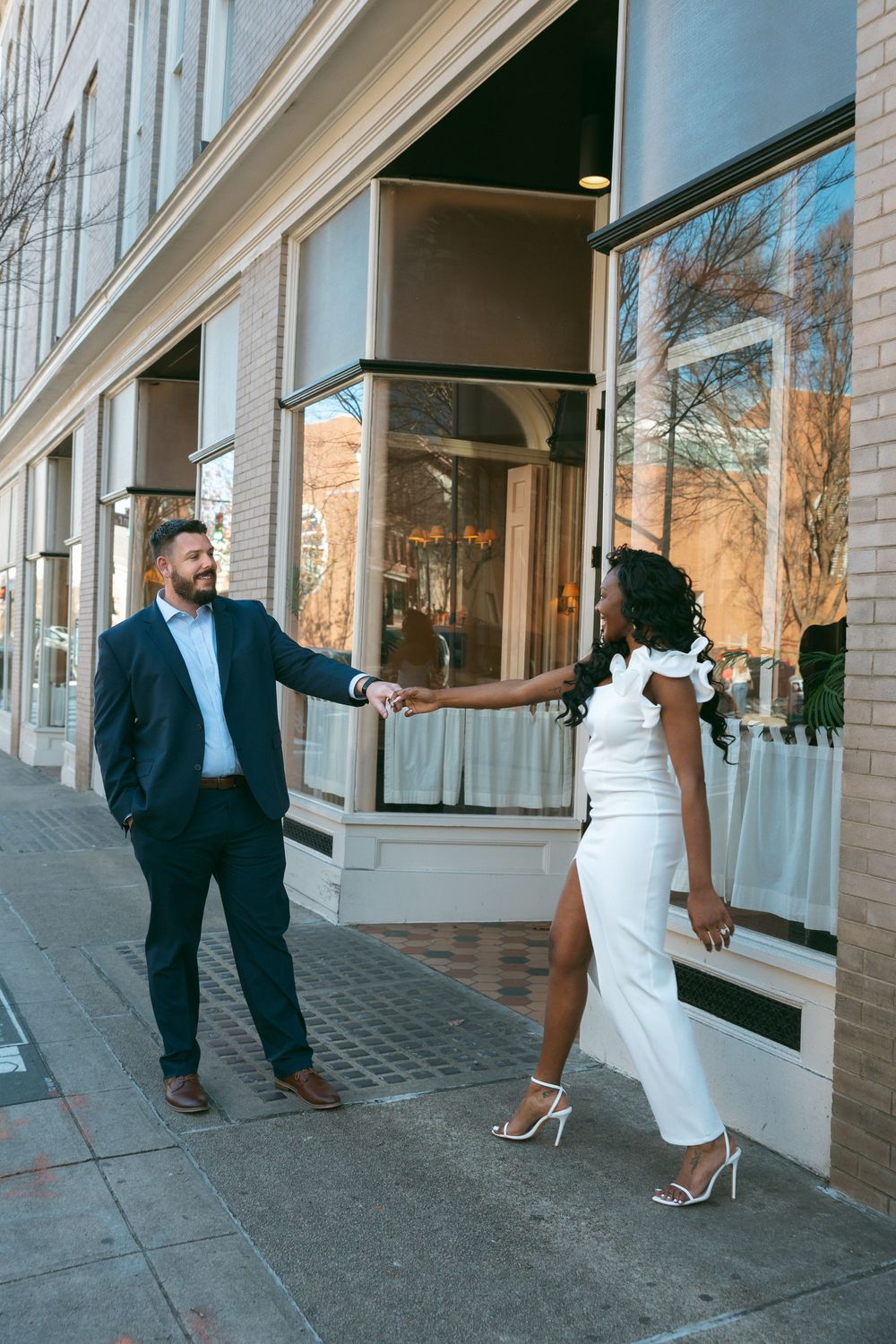 A couple poses playfully in front of a modern building with large windows during a photoshoot.