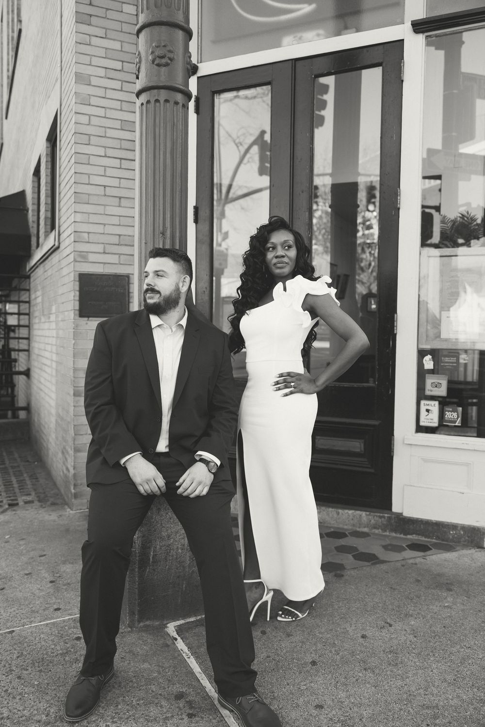 A stylish couple poses in black and white against a brick storefront with architectural columns.