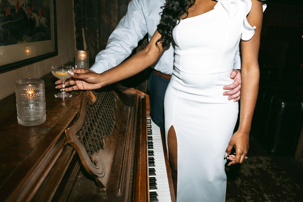 A person in a white dress holds a glass while standing at a bar counter in a dimly lit setting.
