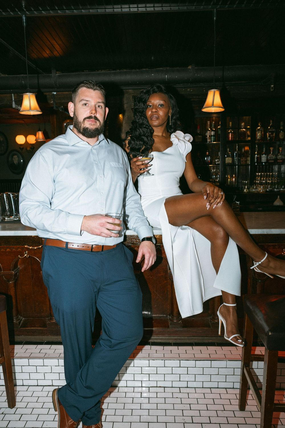 A couple poses casually at a bar with brick walls and ambient lighting.