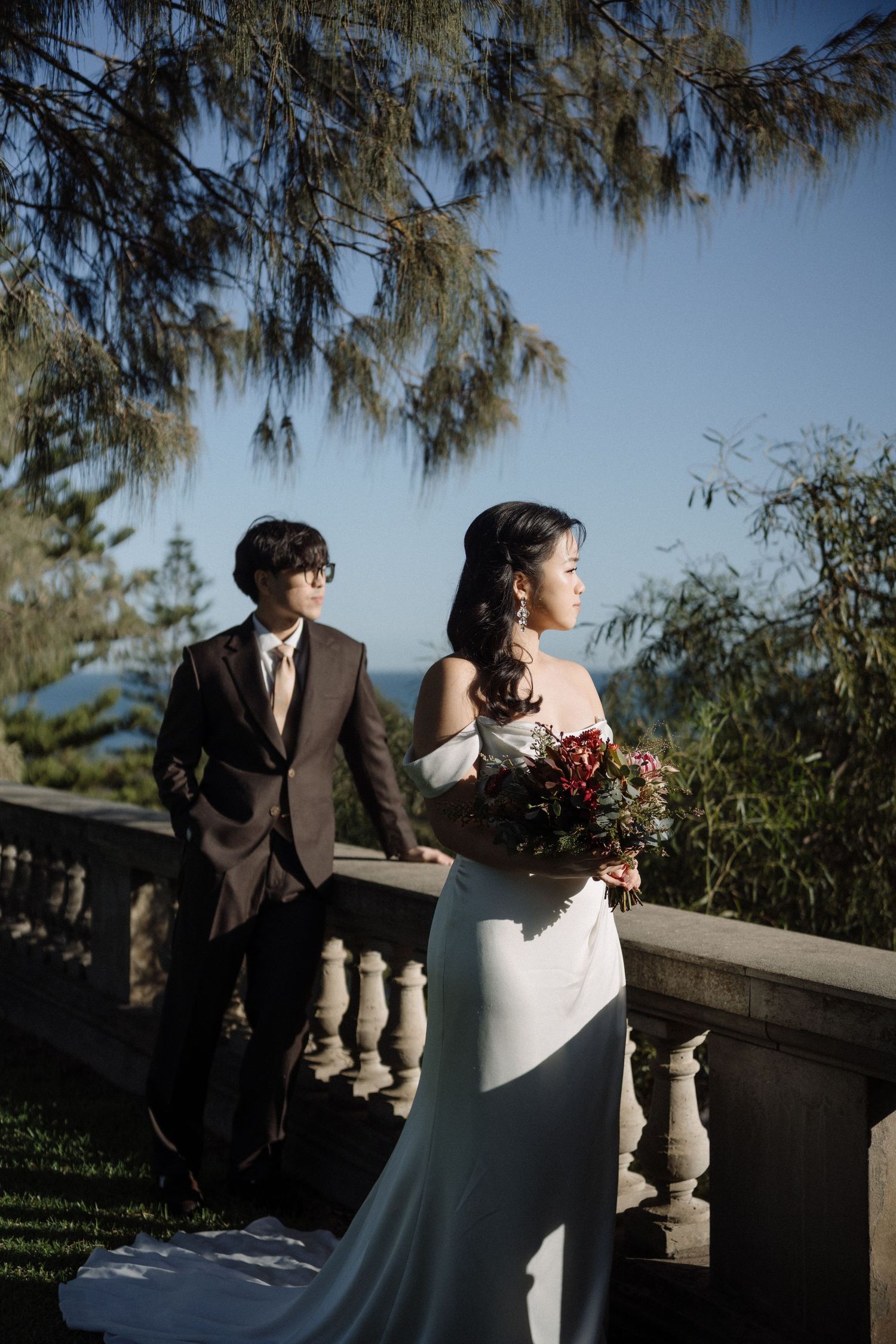 Bride in white off-shoulder gown holds bouquet beside groom in dark suit on stone balcony overlooking ocean.