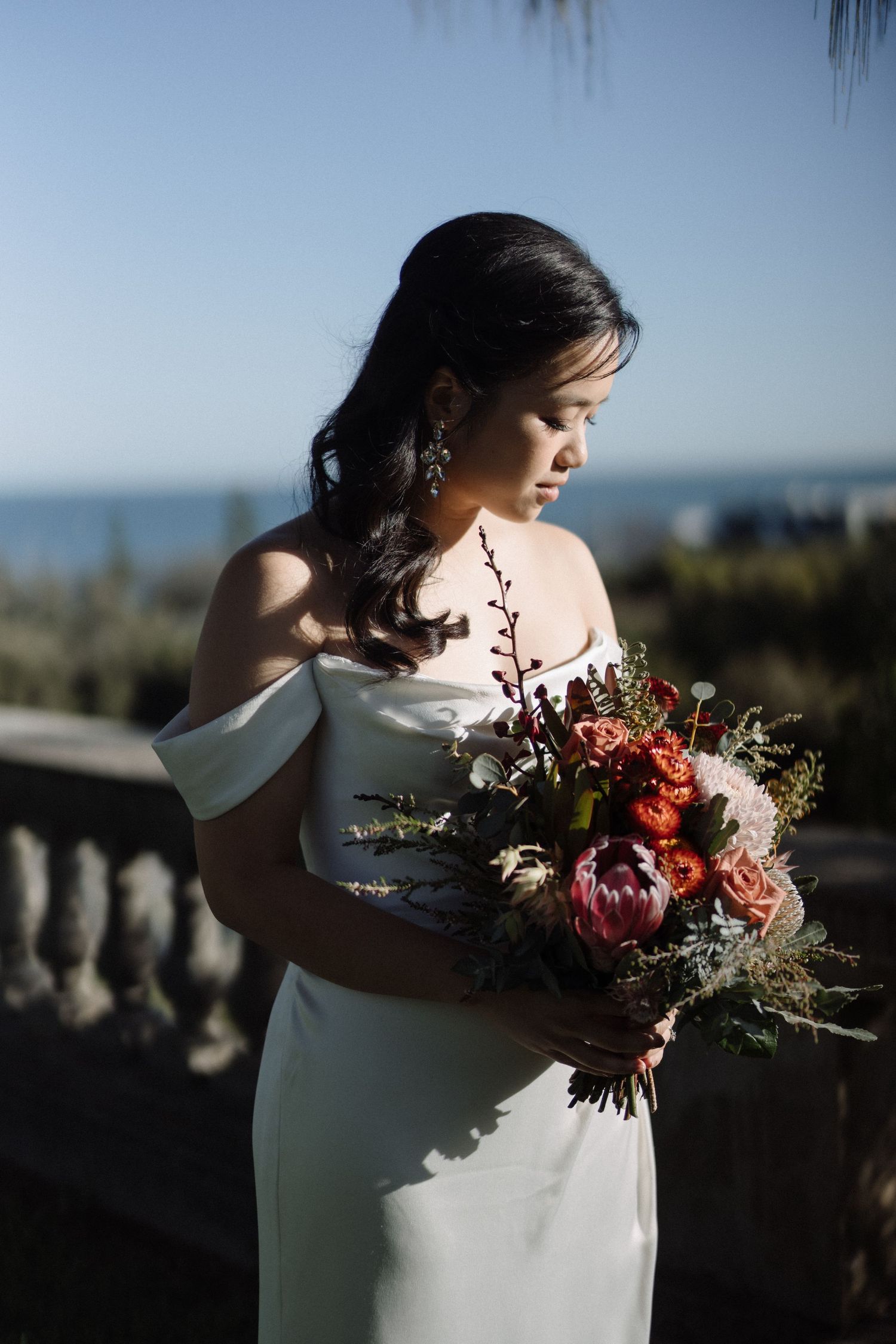 Asian bride in off-shoulder white gown holds vibrant red and pink bouquet with ocean backdrop.