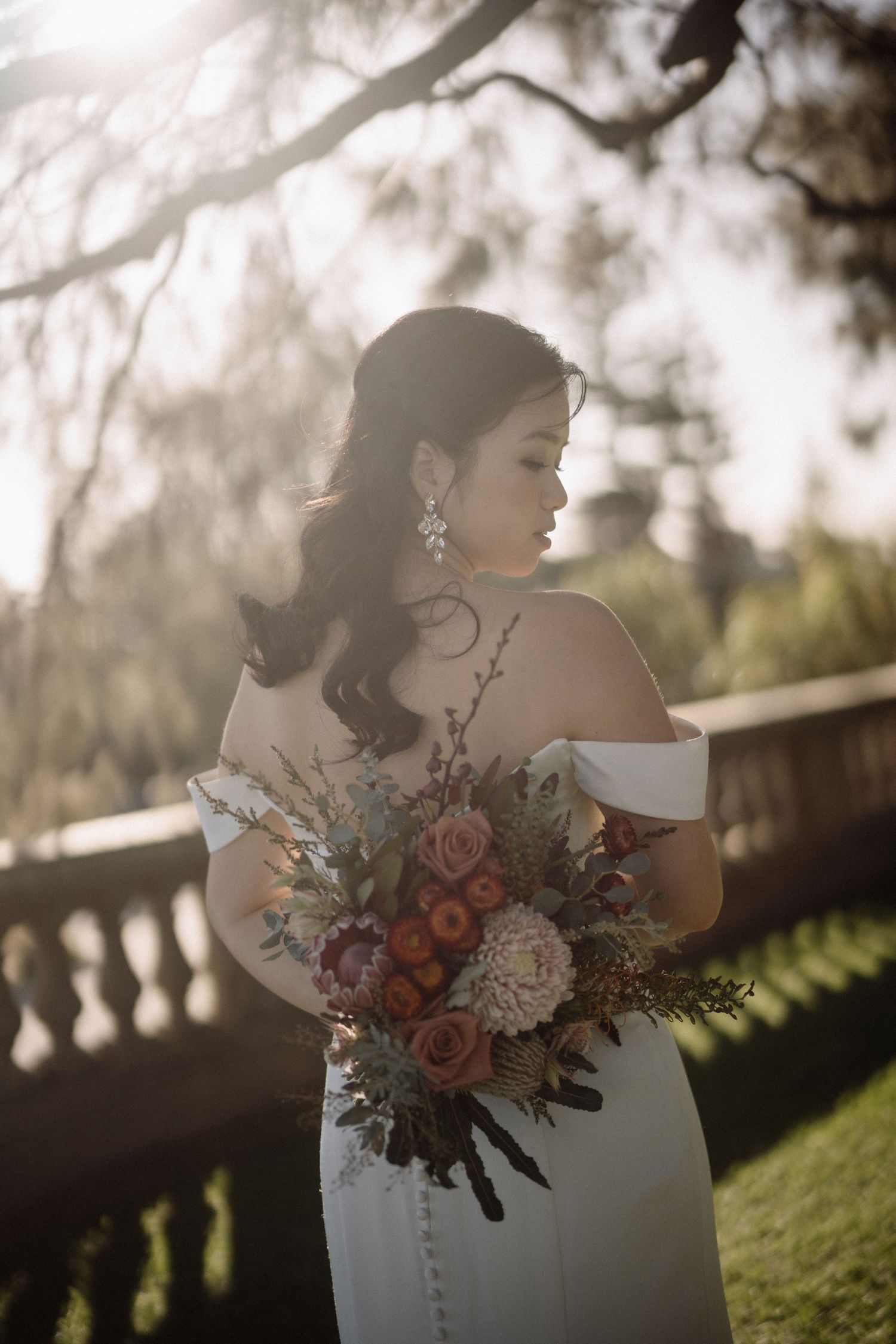 Bride in white off-shoulder gown holds colorful bouquet in golden sunlight with stone balustrade behind.