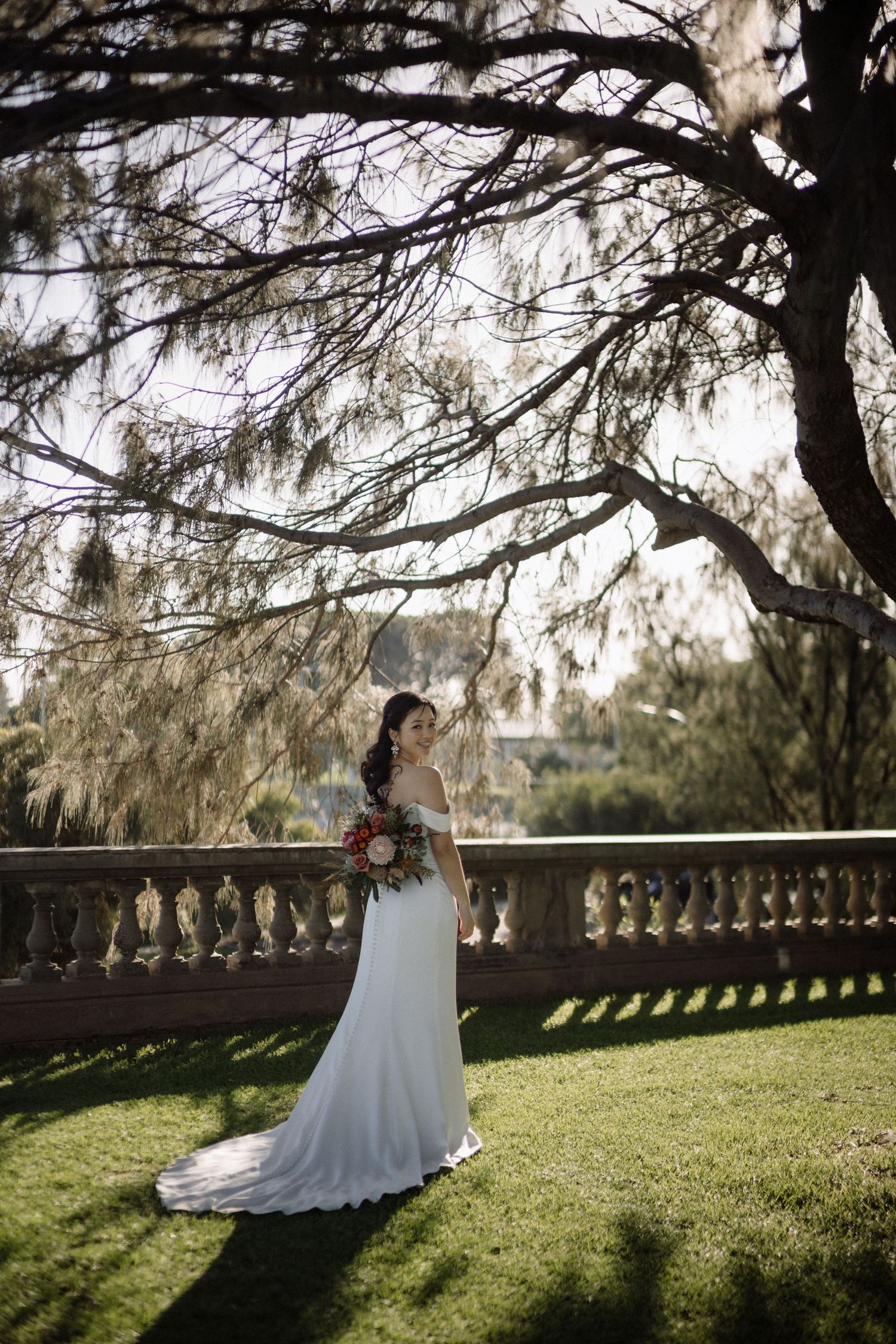 Bride in white off-shoulder gown with train holds bouquet standing by stone balustrade under large tree.