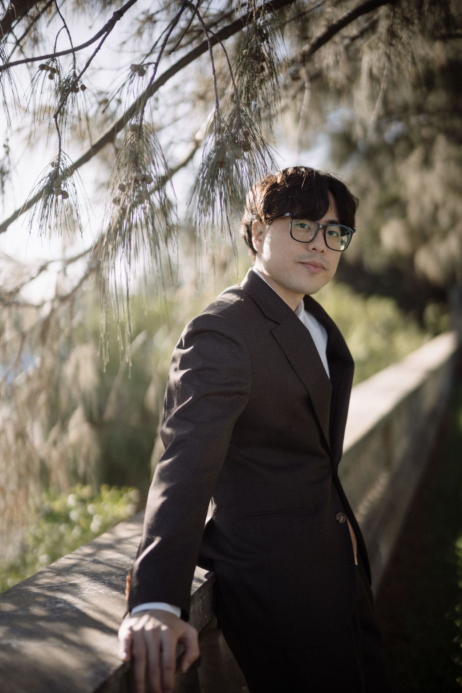 Young man in dark suit and glasses leans on stone railing under pine tree branches outdoors.