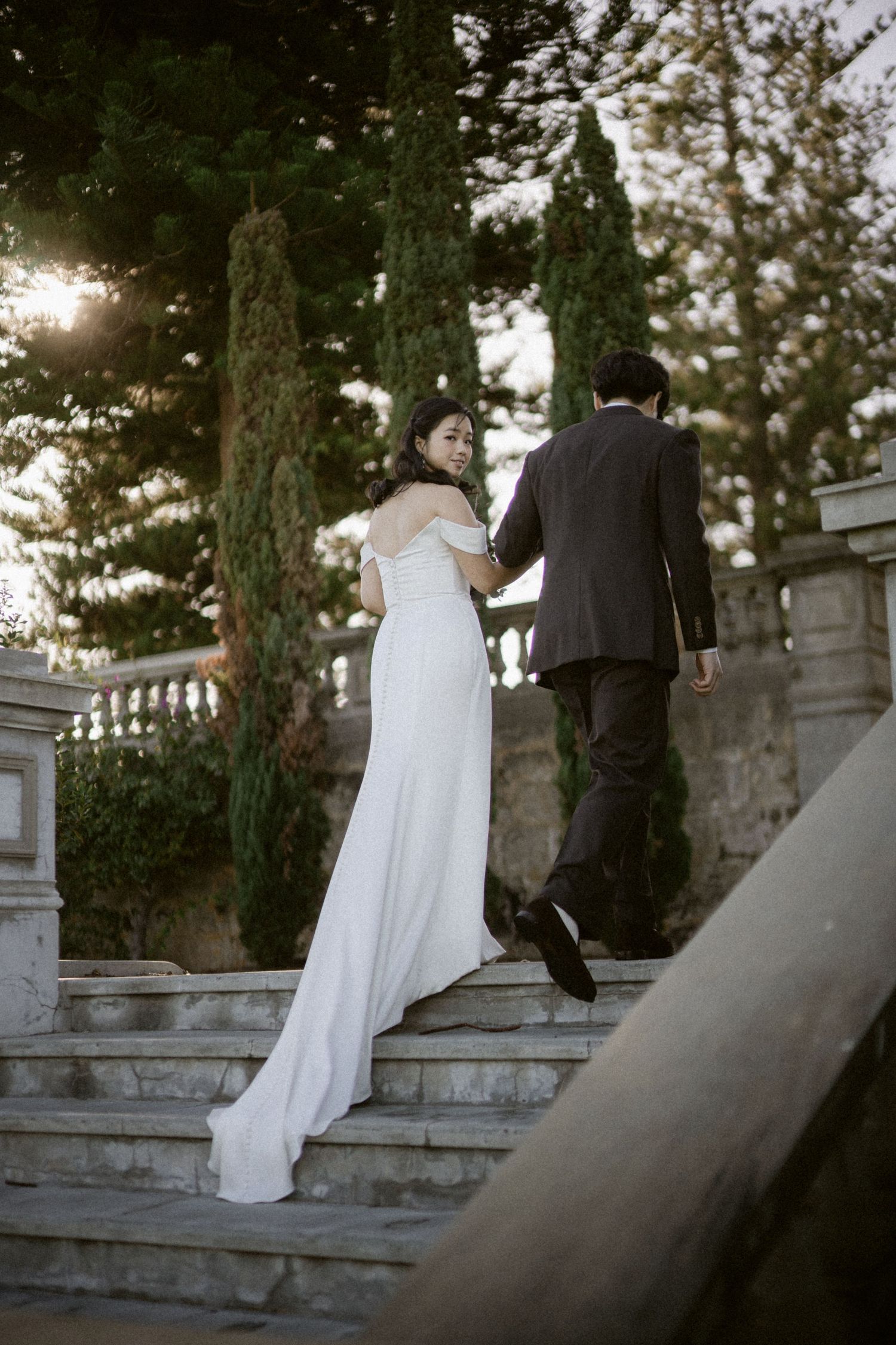 Bride in white gown looks back smiling as groom leads her up stone steps among tall trees.