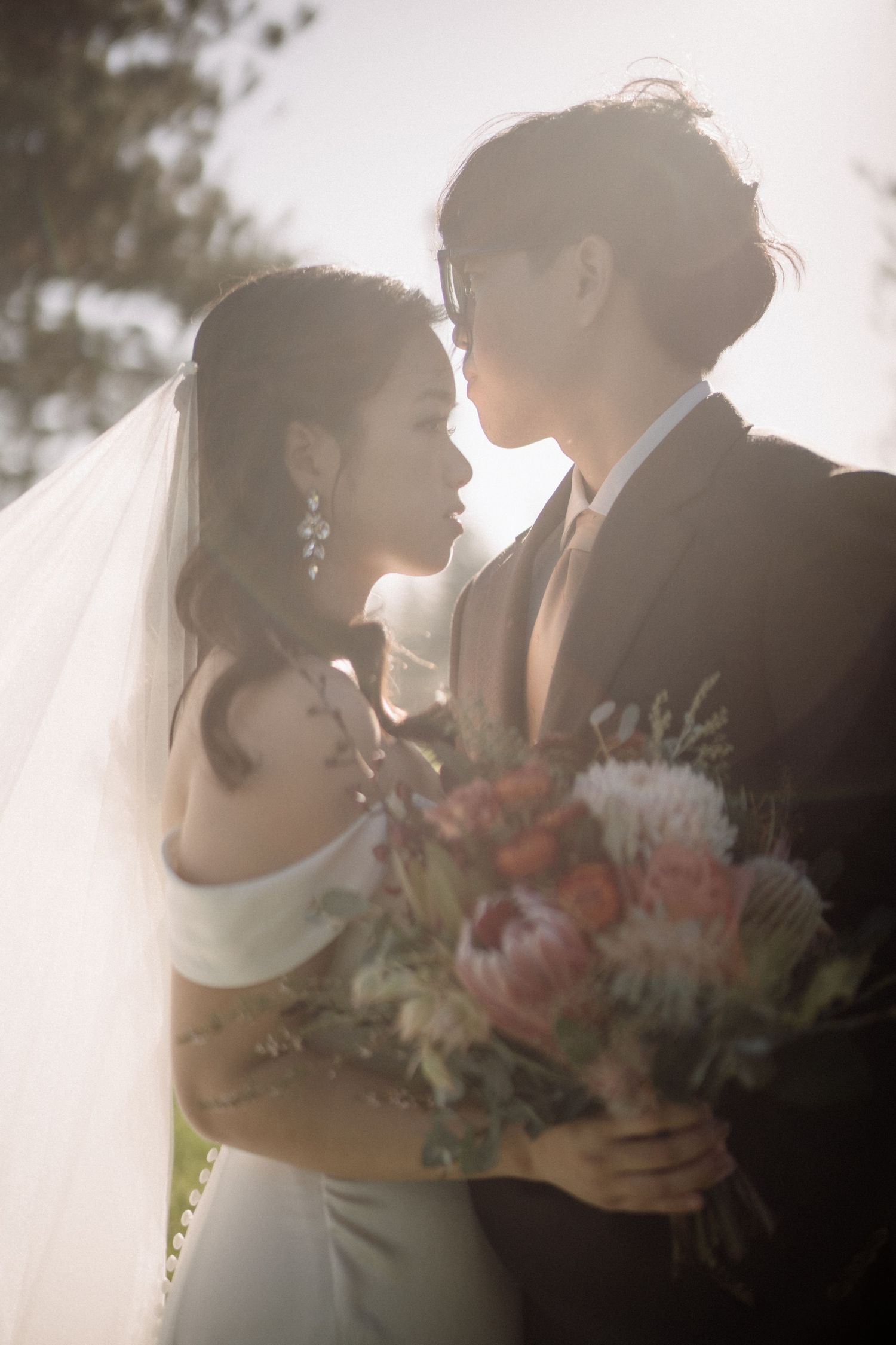 Asian bride and groom lean in for kiss backlit by golden sunlight holding colorful wedding bouquet.