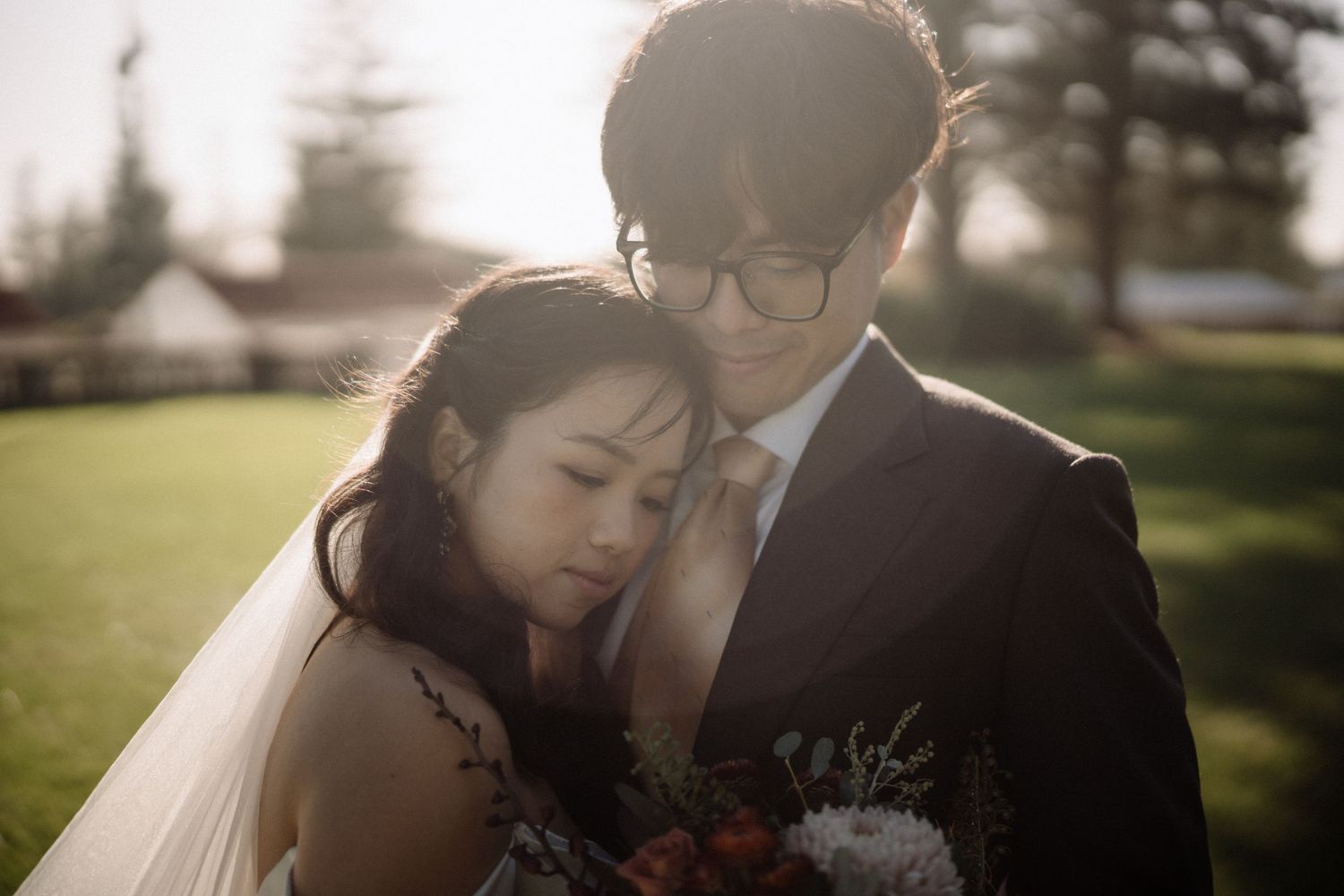 Intimate close-up of bride leaning on groom's chest in warm golden backlight, holding floral bouquet.