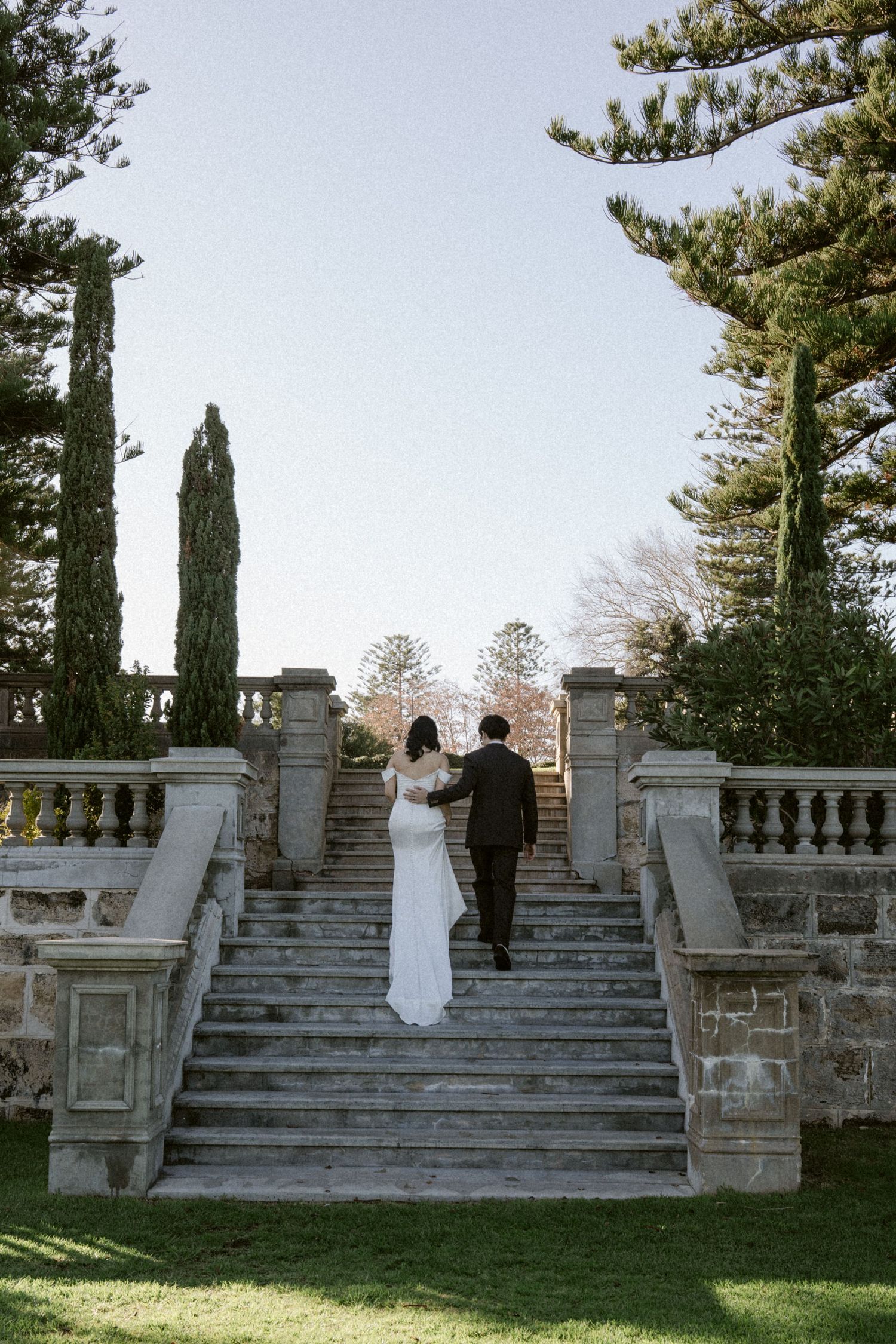 Bride and groom walking up elegant stone staircase in garden.