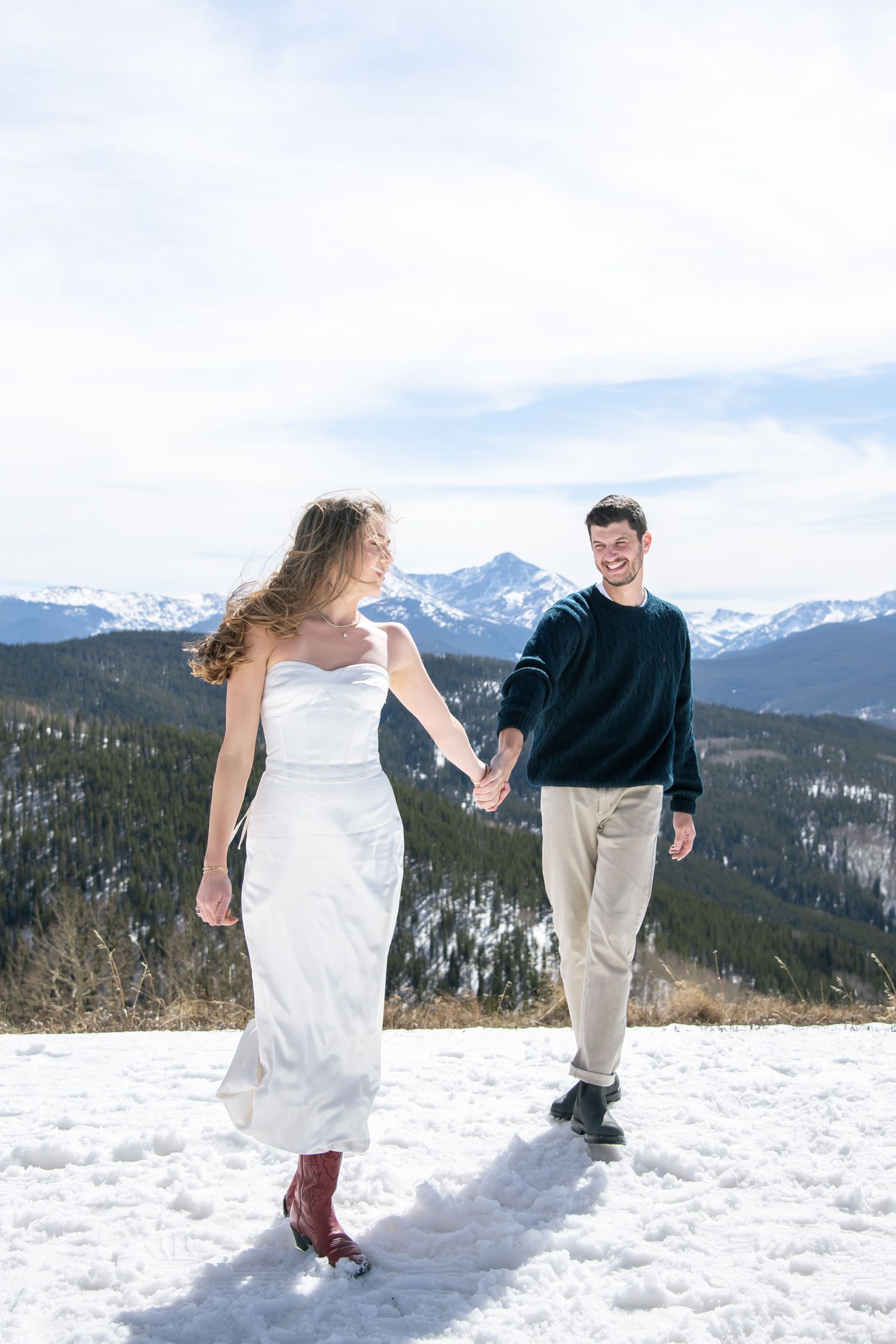 Couple holding hands walking on snowy mountain top with chic red boots and cute outfits for Vail Colorado engagement photos.