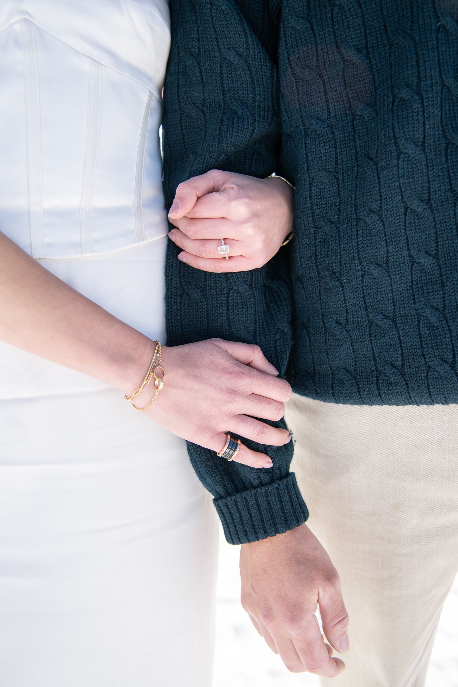 Couple holding hands, woman wearing gold bracelet and ring, man in dark cable-knit sweater, close-up of intertwined arms.