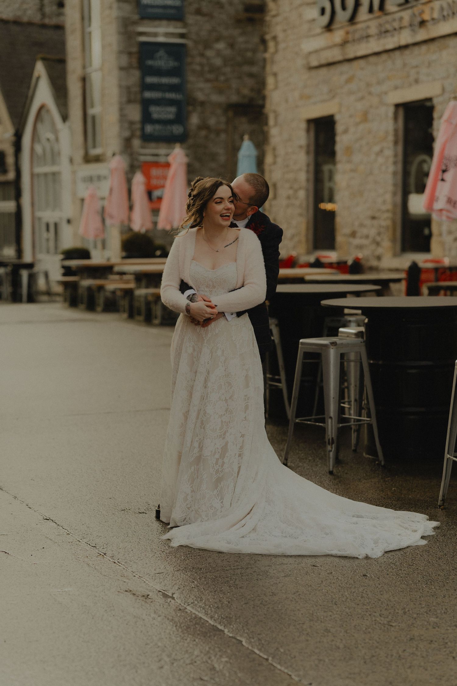 Bride and groom share a romantic embrace in a lace wedding gown and black suit on a cobblestone street.
