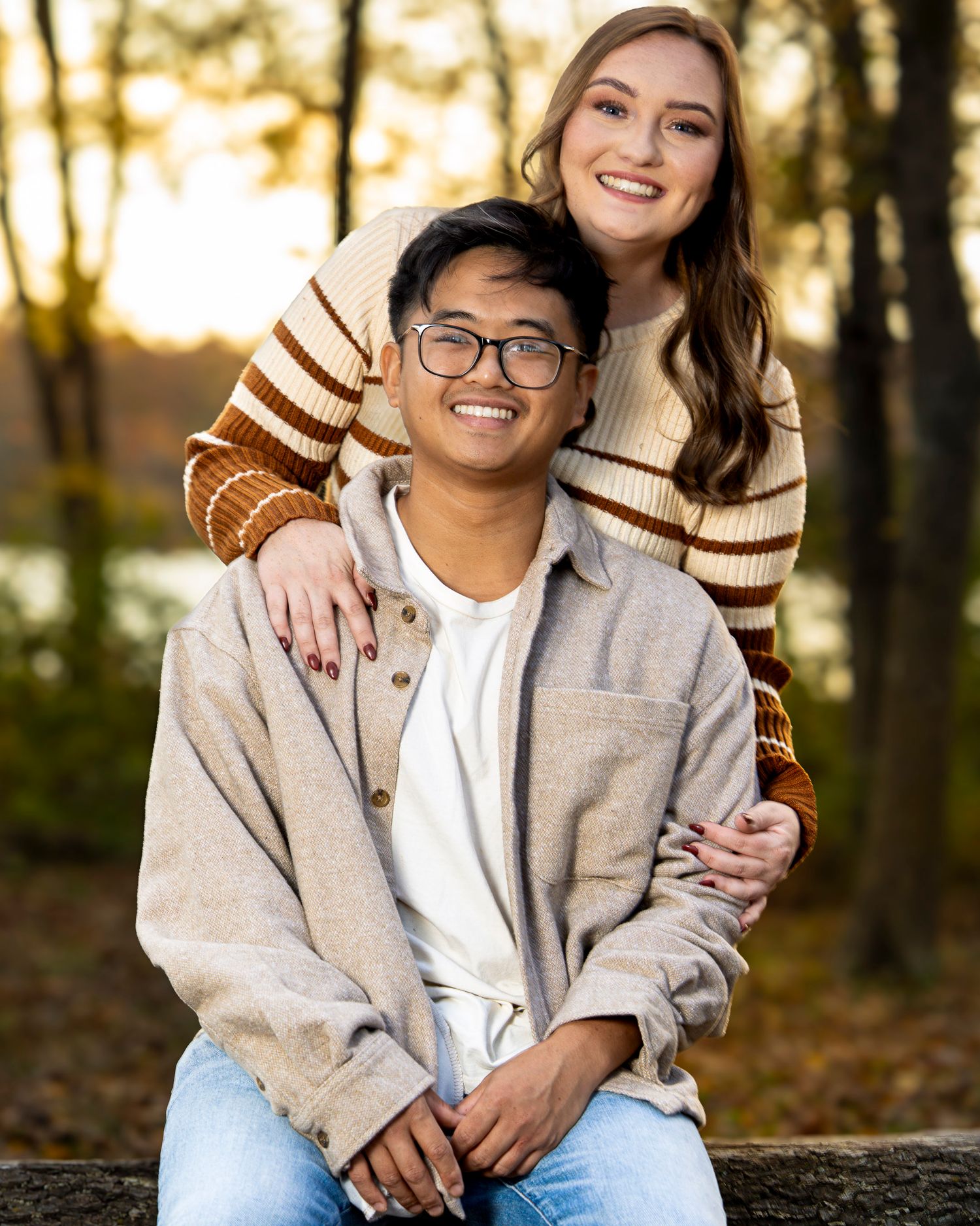 A couple sits together on a fallen tree trunk during golden hour in an autumn forest setting.