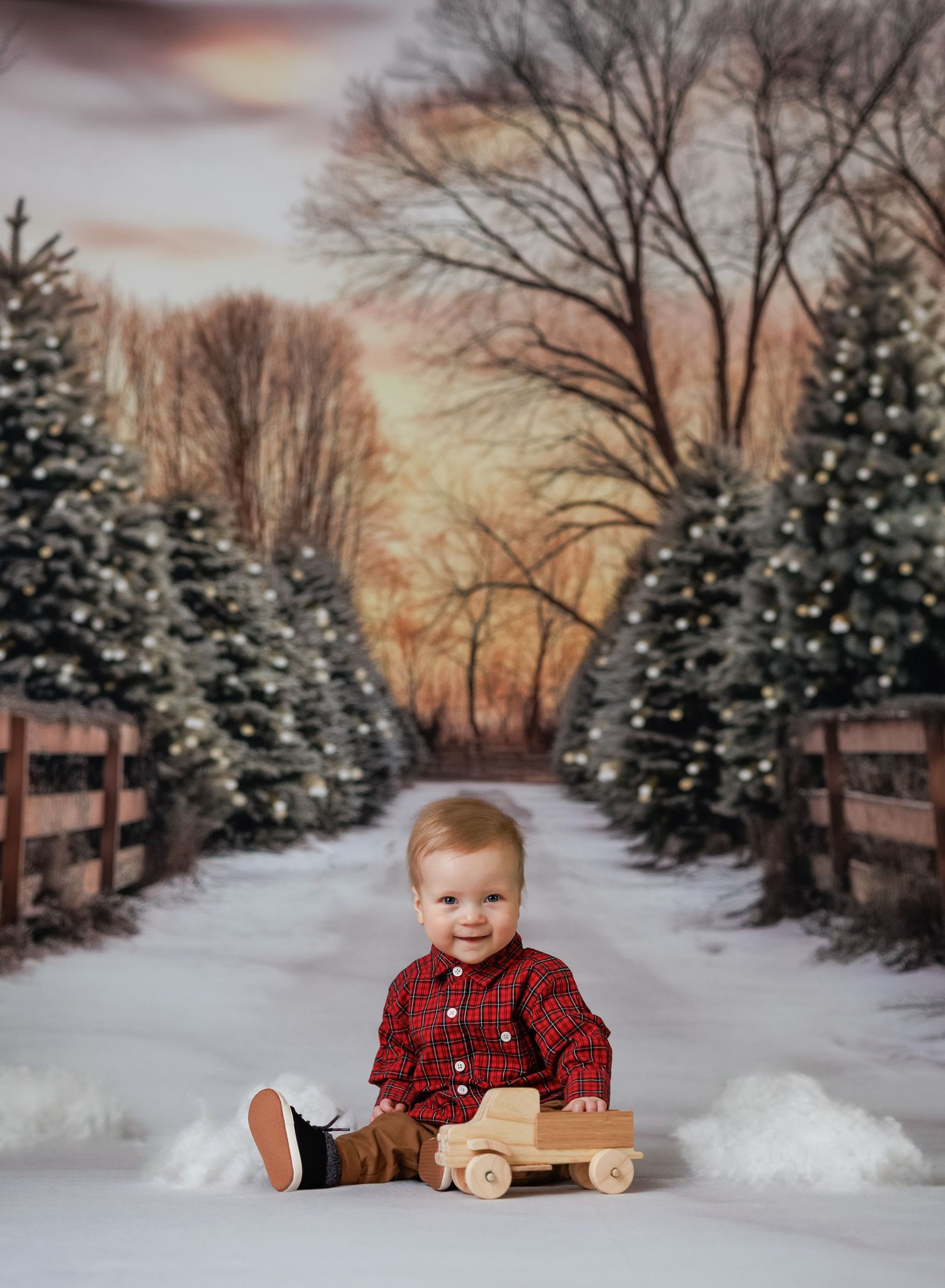 Smiling toddler in red plaid shirt sits with wooden toy truck on snowy path lined with lit Christmas trees.