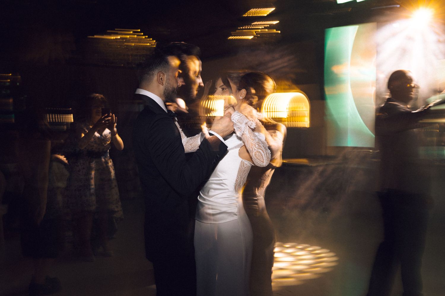 Couple sharing first dance at wedding reception in dimly lit venue with warm lighting and guests in background.