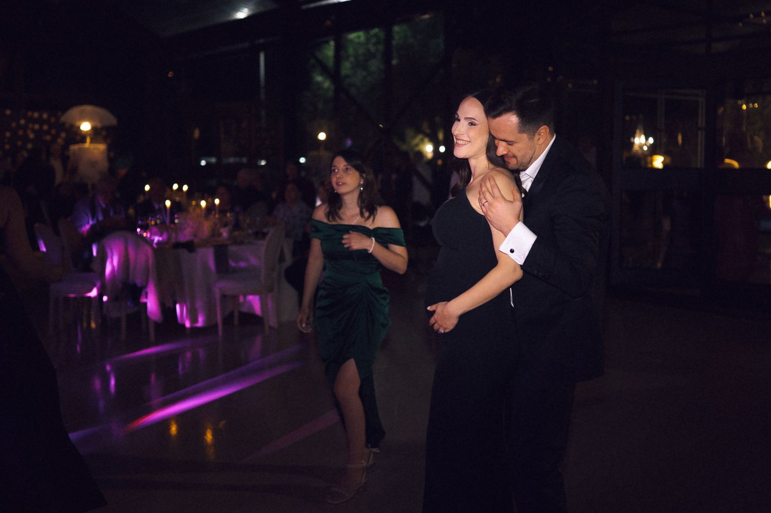 Elegant couple sharing a slow dance at a formal evening event with purple lighting and guests in background.
