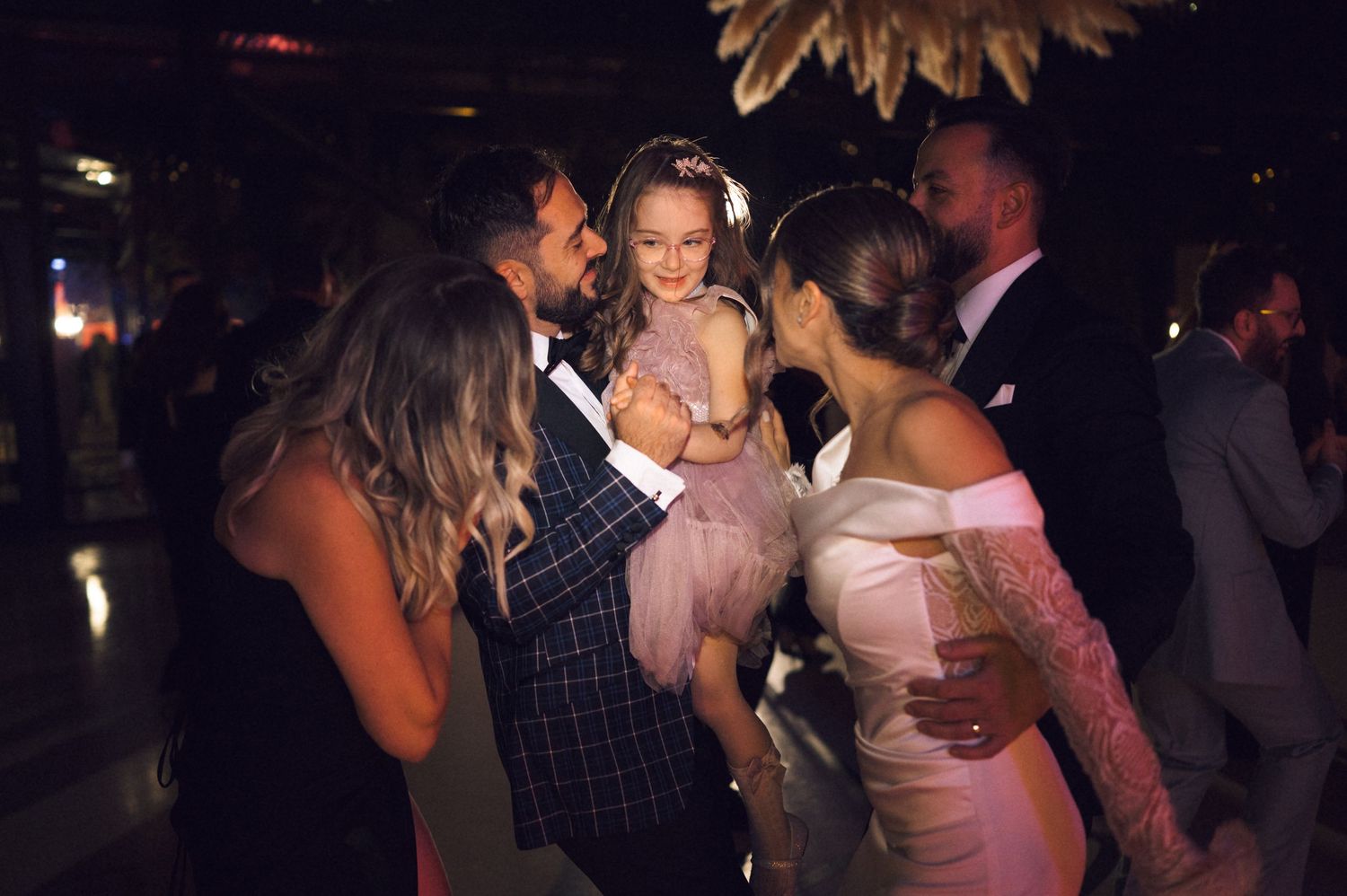 A joyful wedding reception scene with guests dancing, a man holding a young girl in a pink dress, and a bride in white.