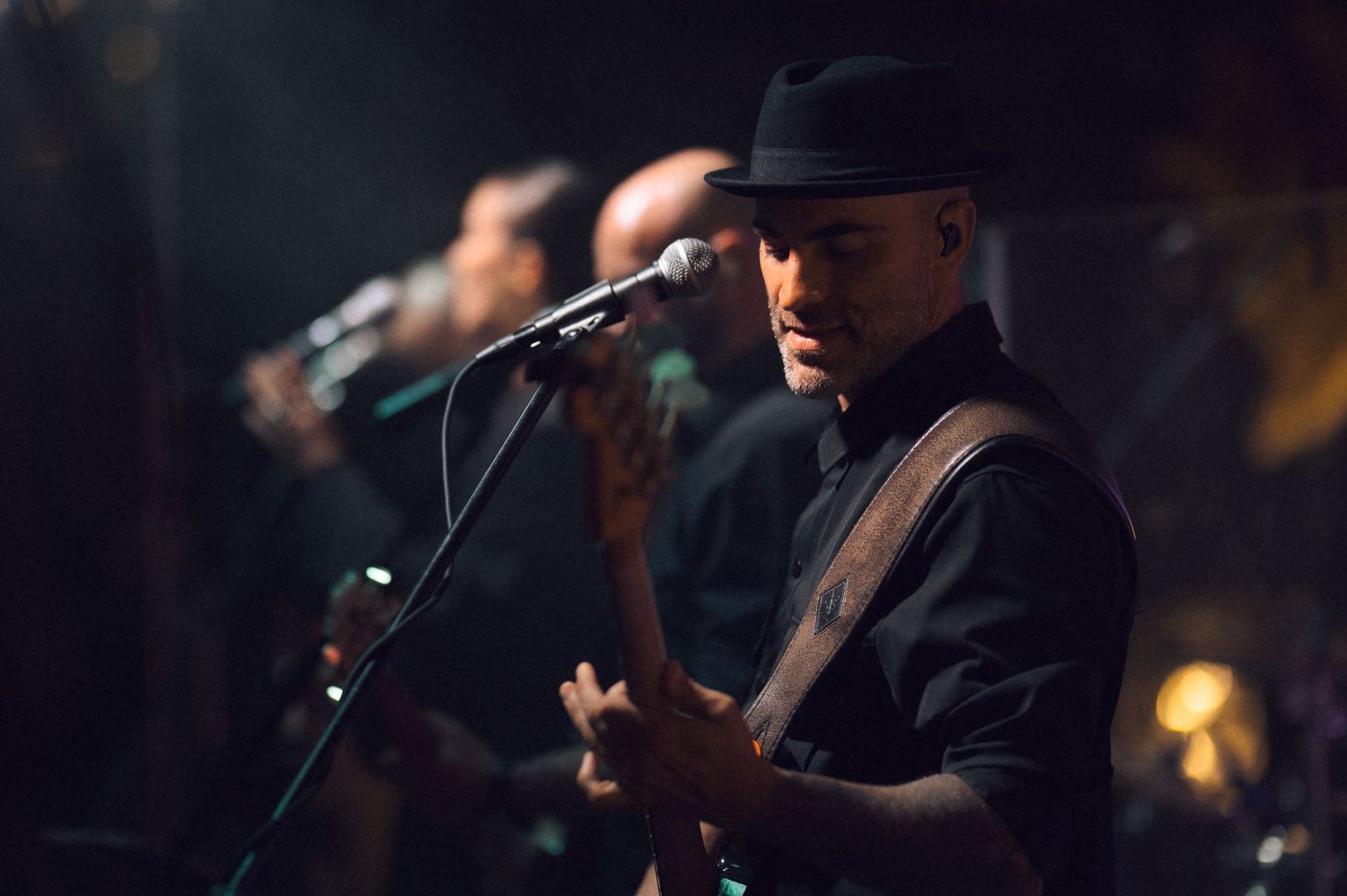 A young Black musician in a black hat performs at a microphone on a dimly lit stage with band members behind him.