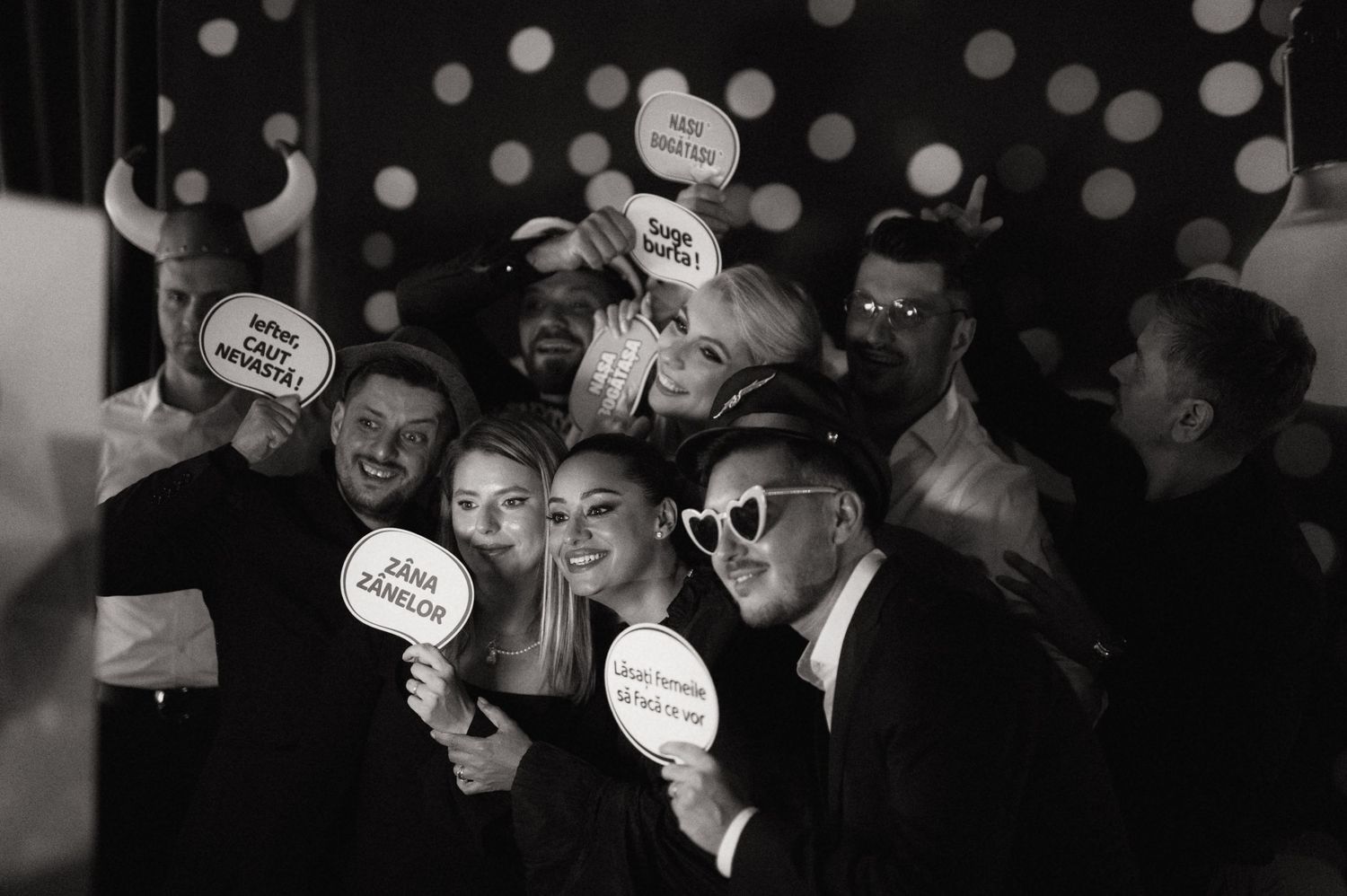 Black and white photo booth shot of a group of friends holding fun signs and props at a party or wedding event.