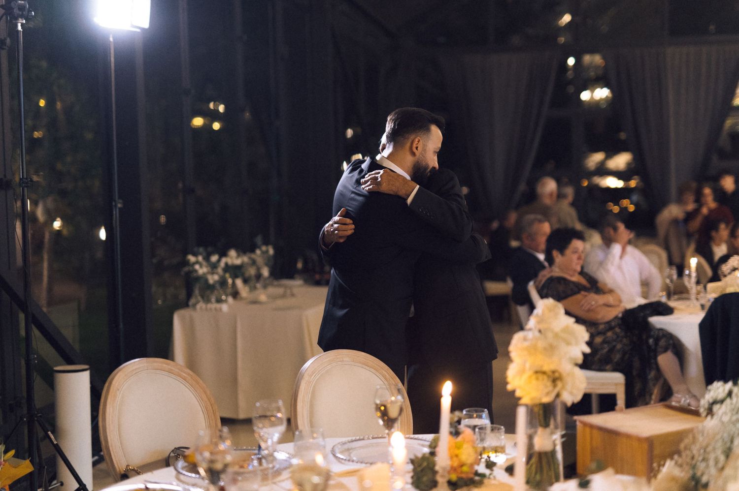 Two grooms share a first dance at an elegant candlelit wedding reception with floral centerpieces.