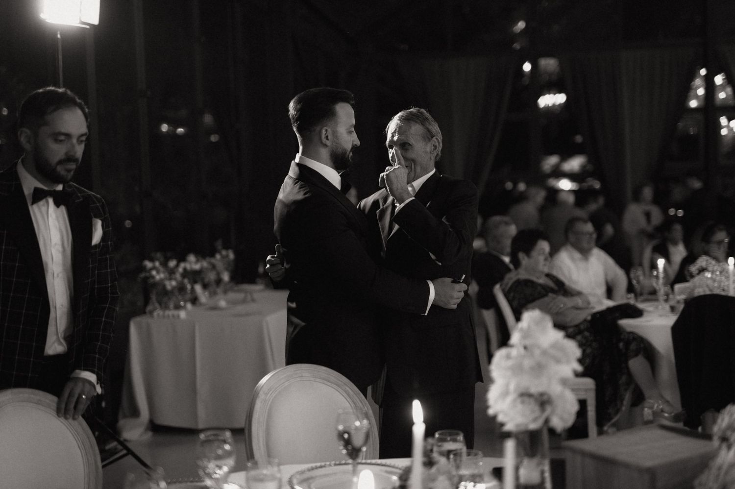 Black and white wedding reception photo of two men sharing a dance moment under elegant candlelit venue lighting.
