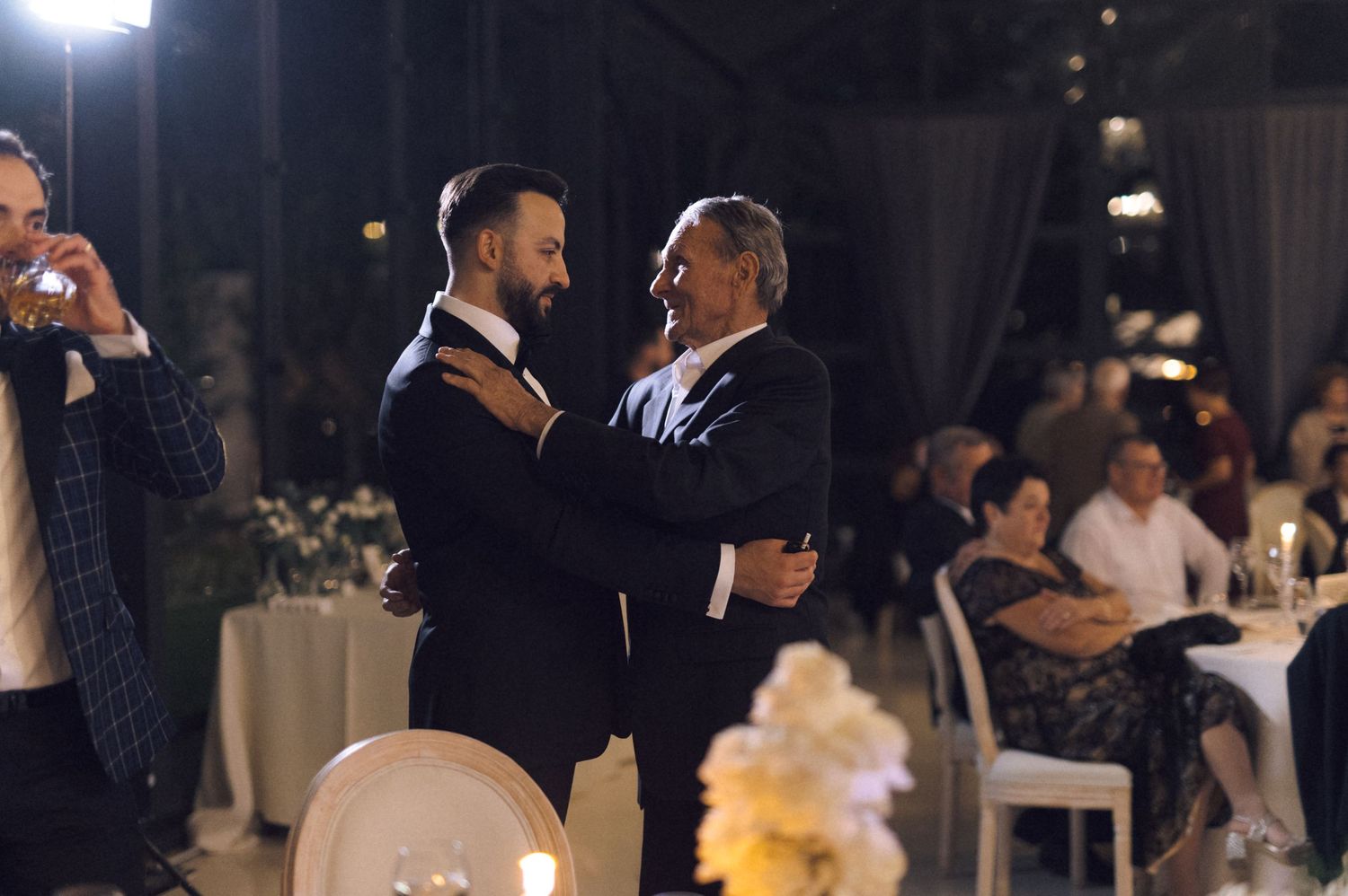 Two men in tuxedos share a dance at an elegant evening wedding reception with guests seated in background.