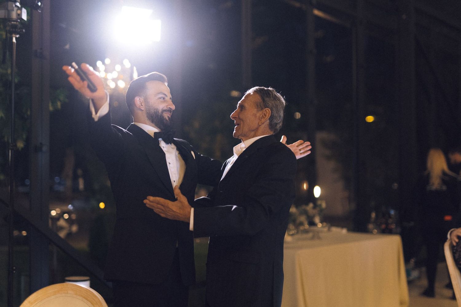 Two men in tuxedos share a joyful dance at an elegant evening reception with warm lighting.