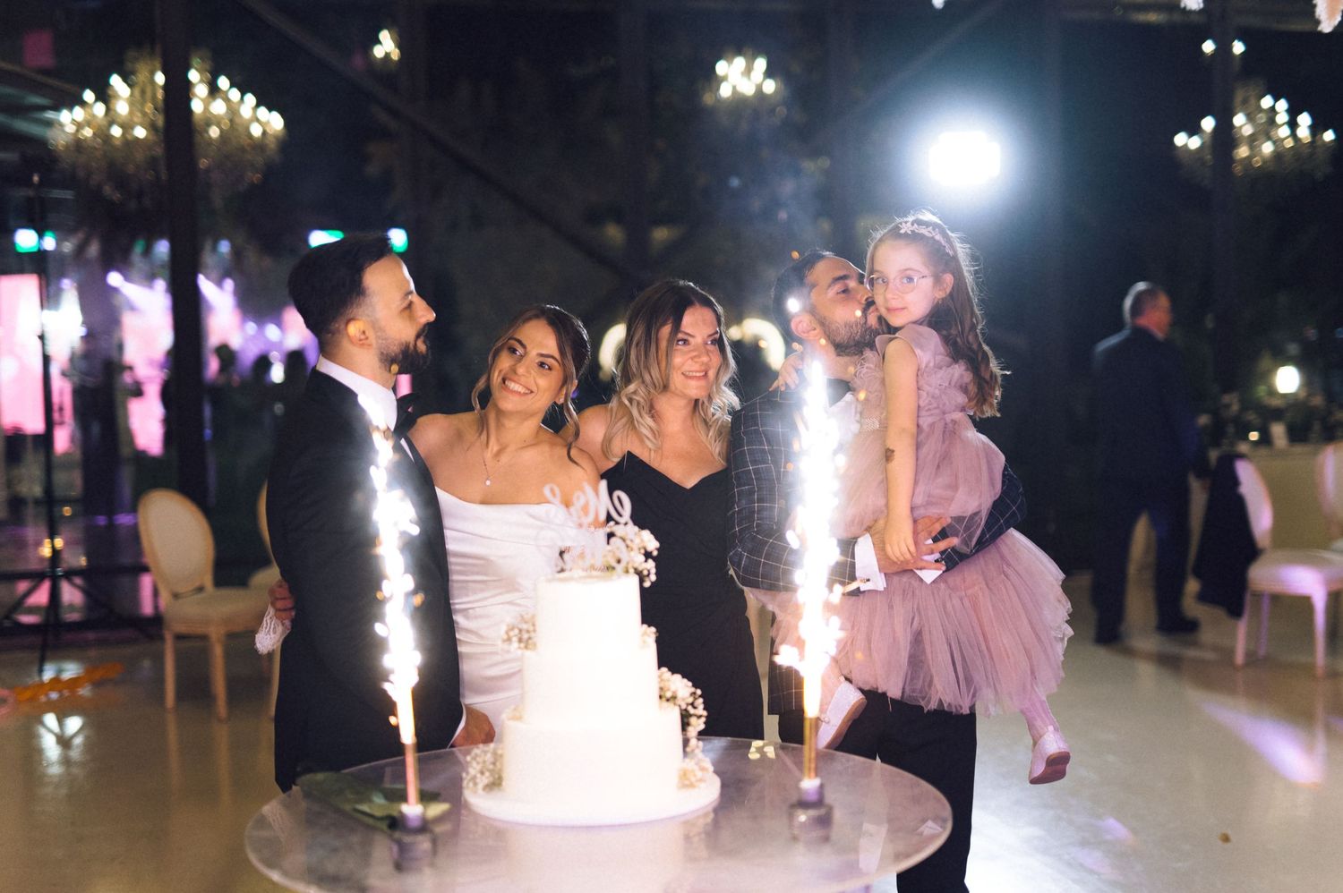 Wedding couple kisses near tiered cake with sparklers, joined by two women and a child in pink dress at elegant reception.