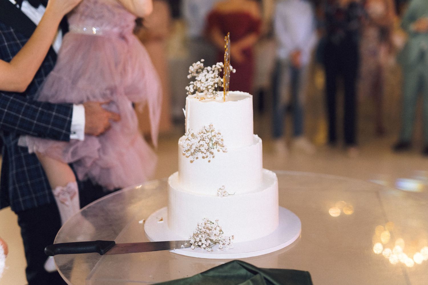 Elegant three-tier white wedding cake with gold topper and baby's breath flowers on a silver stand.