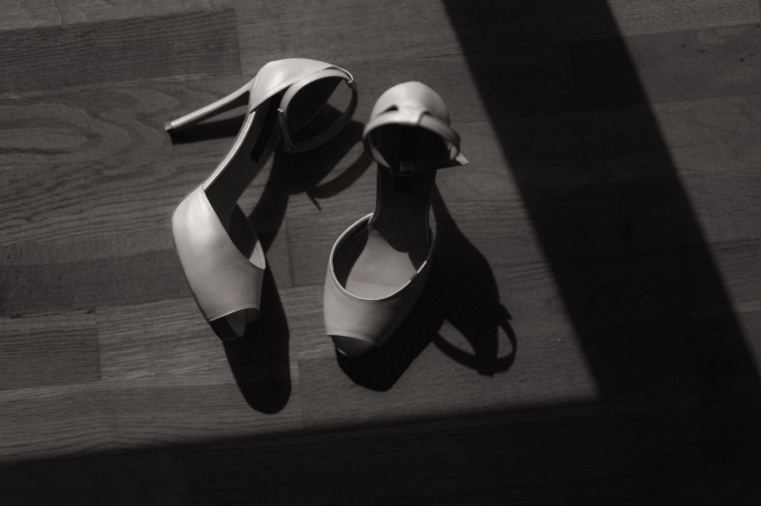 Black and white photo of elegant high-heel sandals with ankle straps resting on a wooden floor.