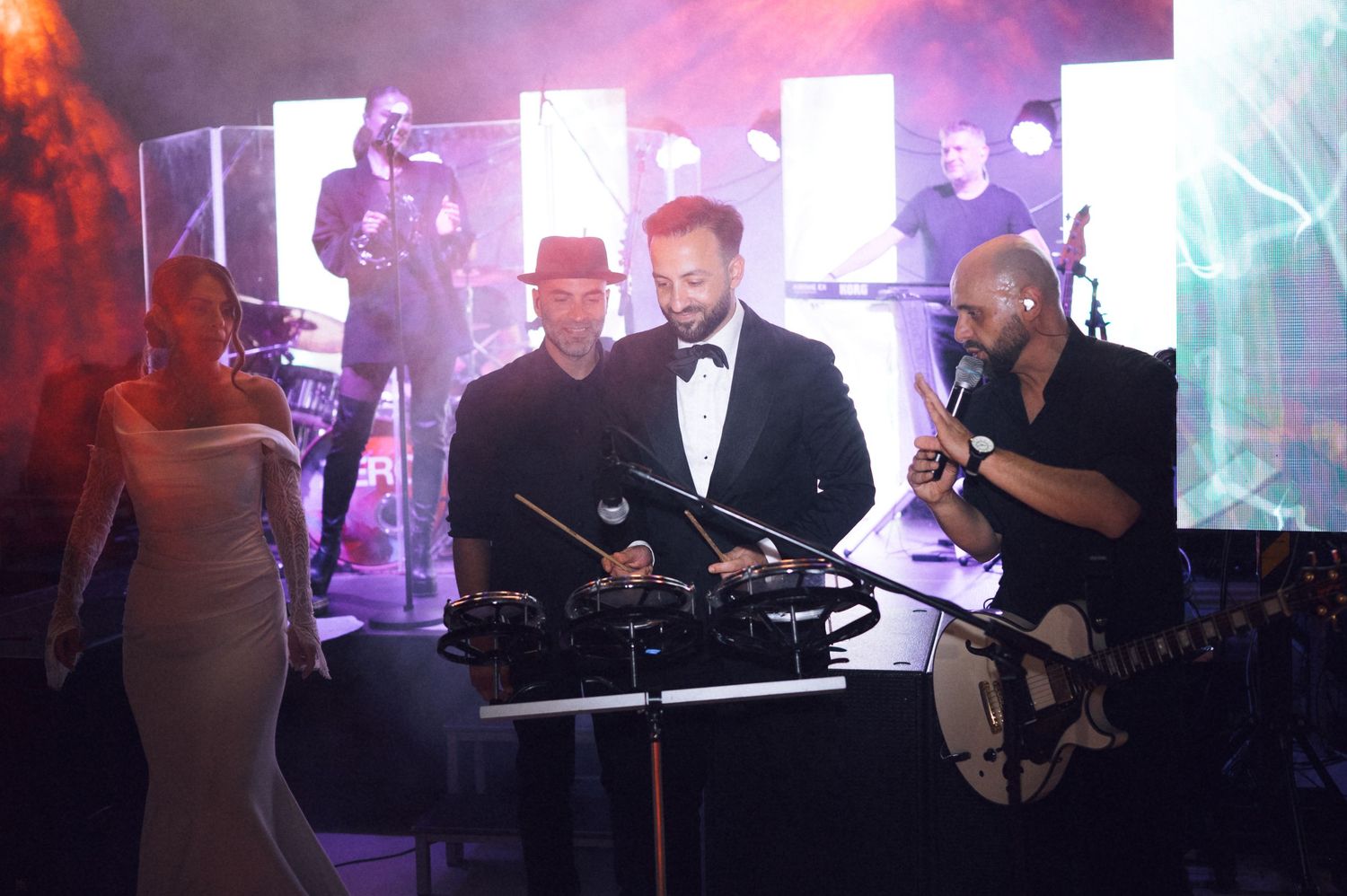 Live band performance on stage with musicians, DJ equipment, and colorful lighting at an evening event.
