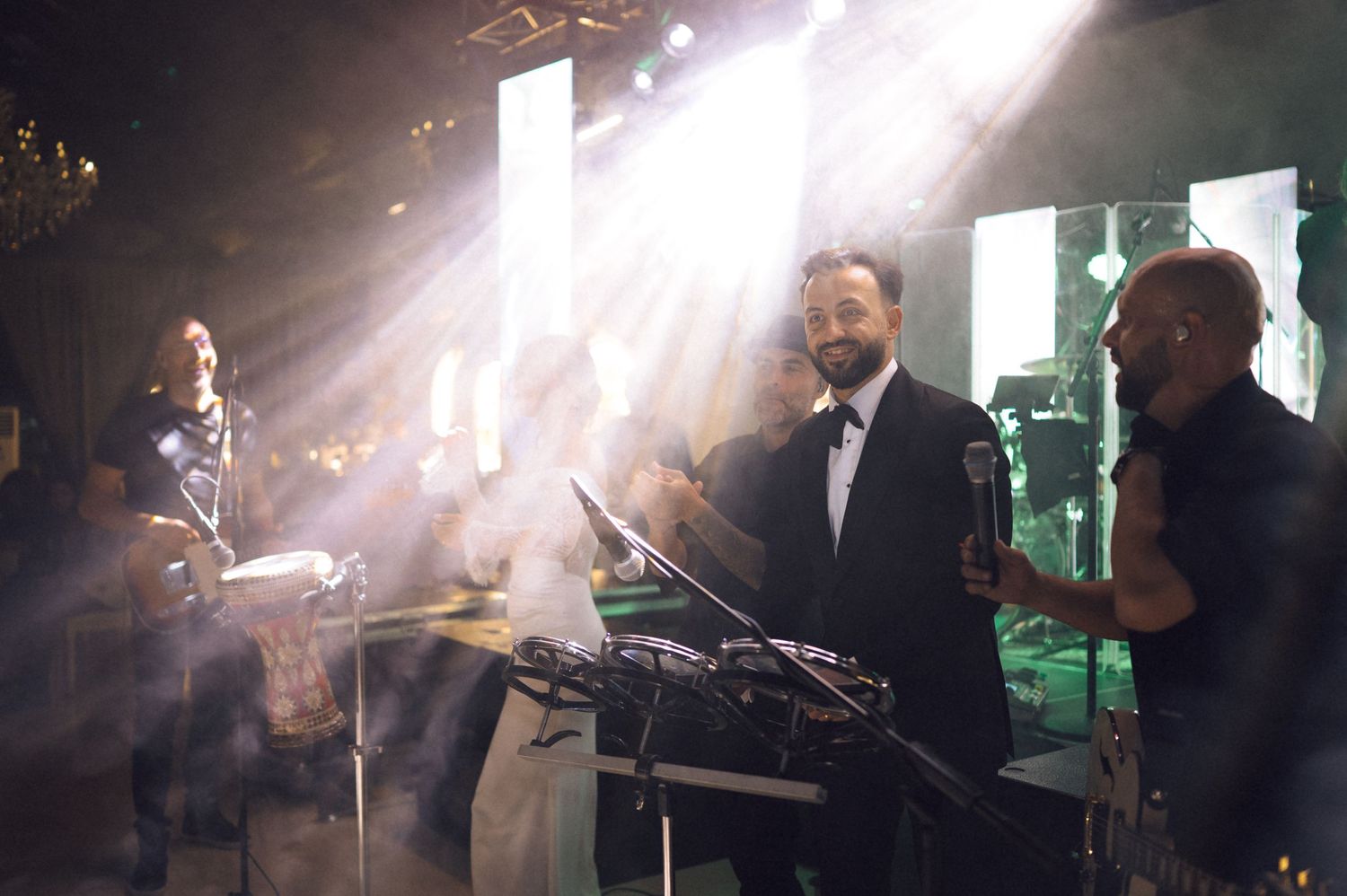A man in a tuxedo smiles on stage with band members under dramatic spotlights at an elegant event.