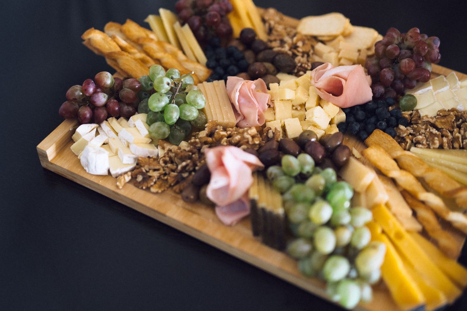 Elegant charcuterie board with assorted cheeses, deli meats, green and red grapes, nuts, and breadsticks on wooden platter.